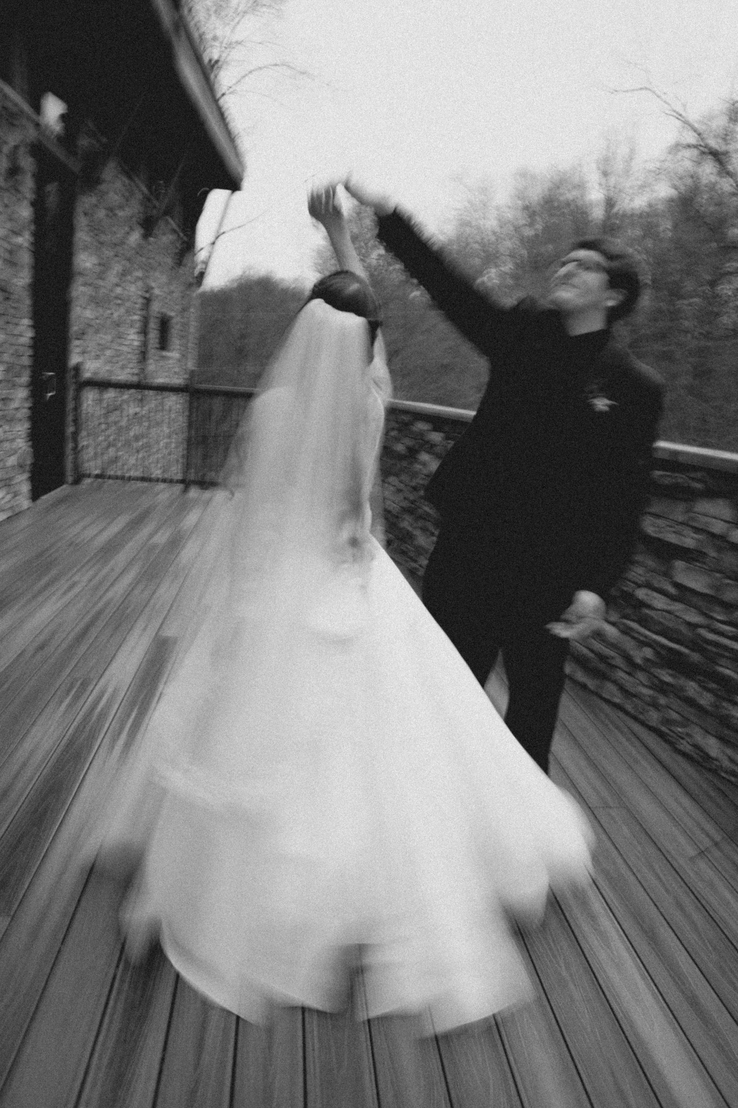 Black and white photo of a bride in a white gown spinning with a groom on a wooden deck during their wedding.