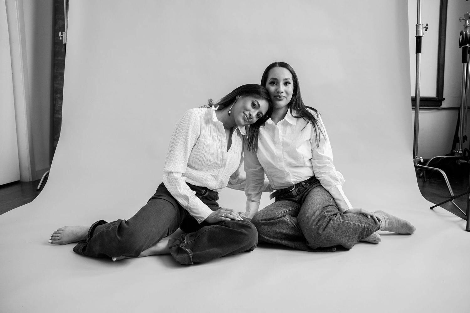 Two women in white shirts and jeans sit together on studio floor, smiling, in a black and white photo.