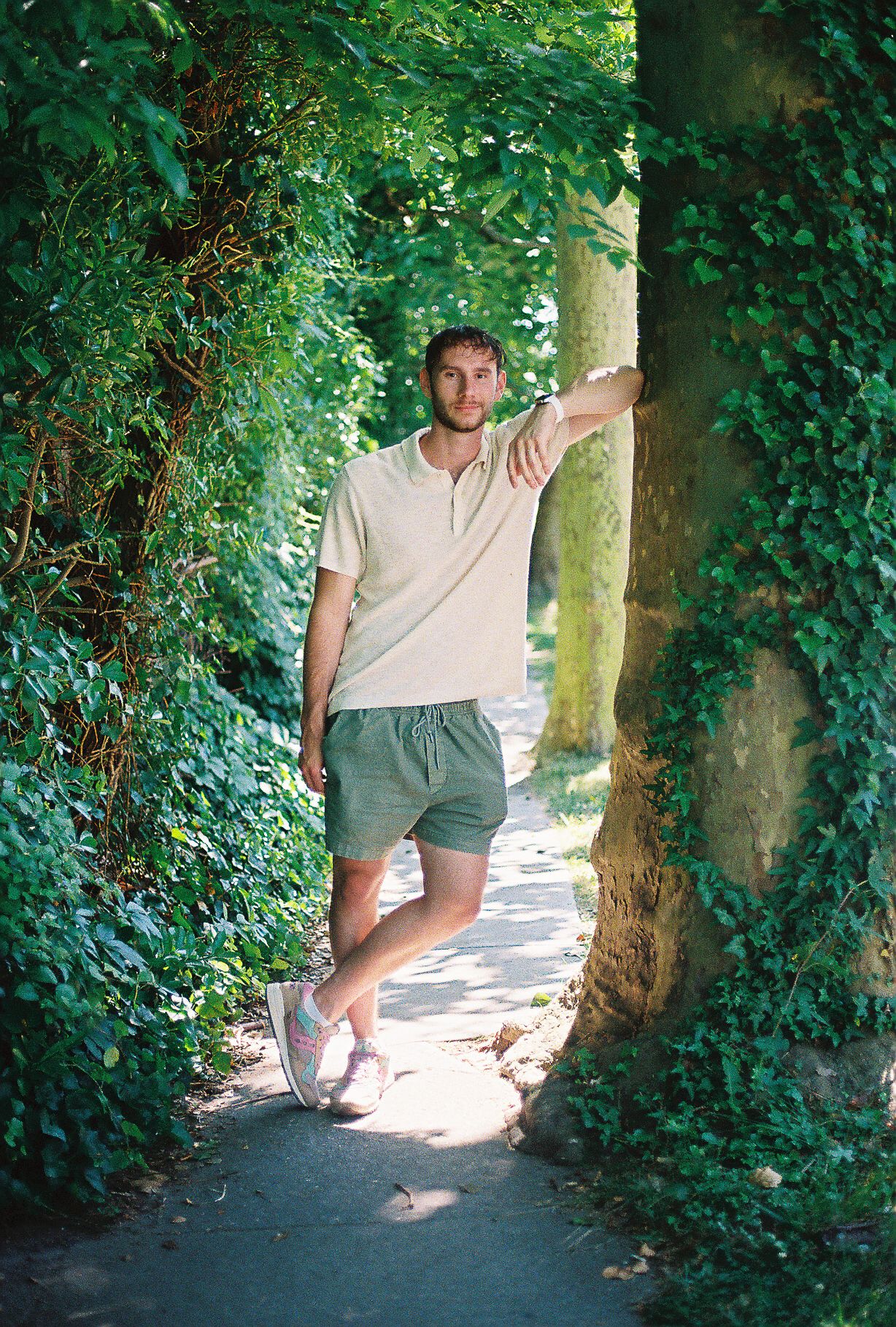 Young man leaning against ivy-covered tree on shaded garden pathway wearing shorts and polo shirt.