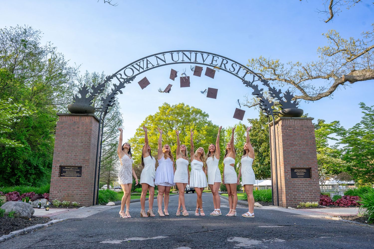 Group of graduates in white dresses tossing caps at Rowan University entrance gate.