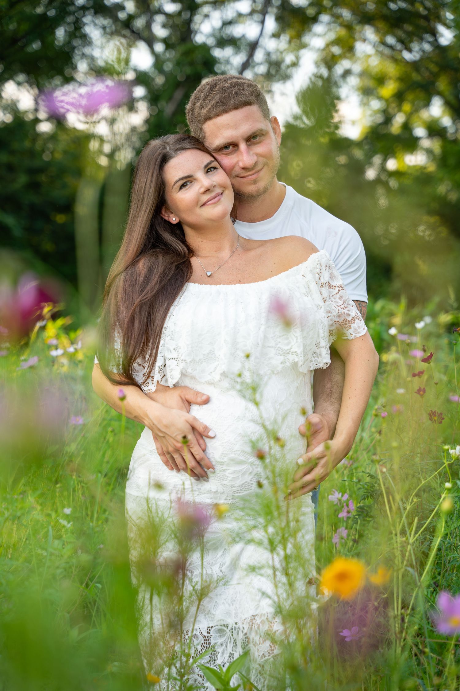 Couple embracing in flower field, woman in white dress with visible baby bump outdoors.