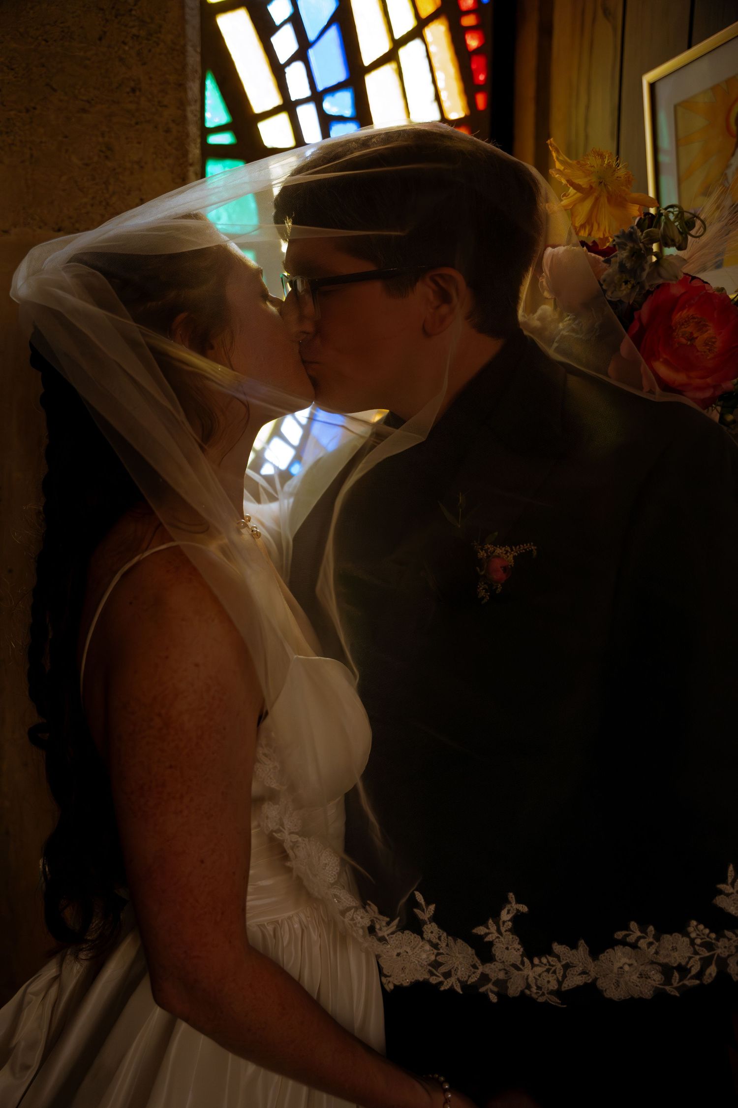 Bride and groom sharing a romantic kiss under a veil in a church with colorful stained glass windows.