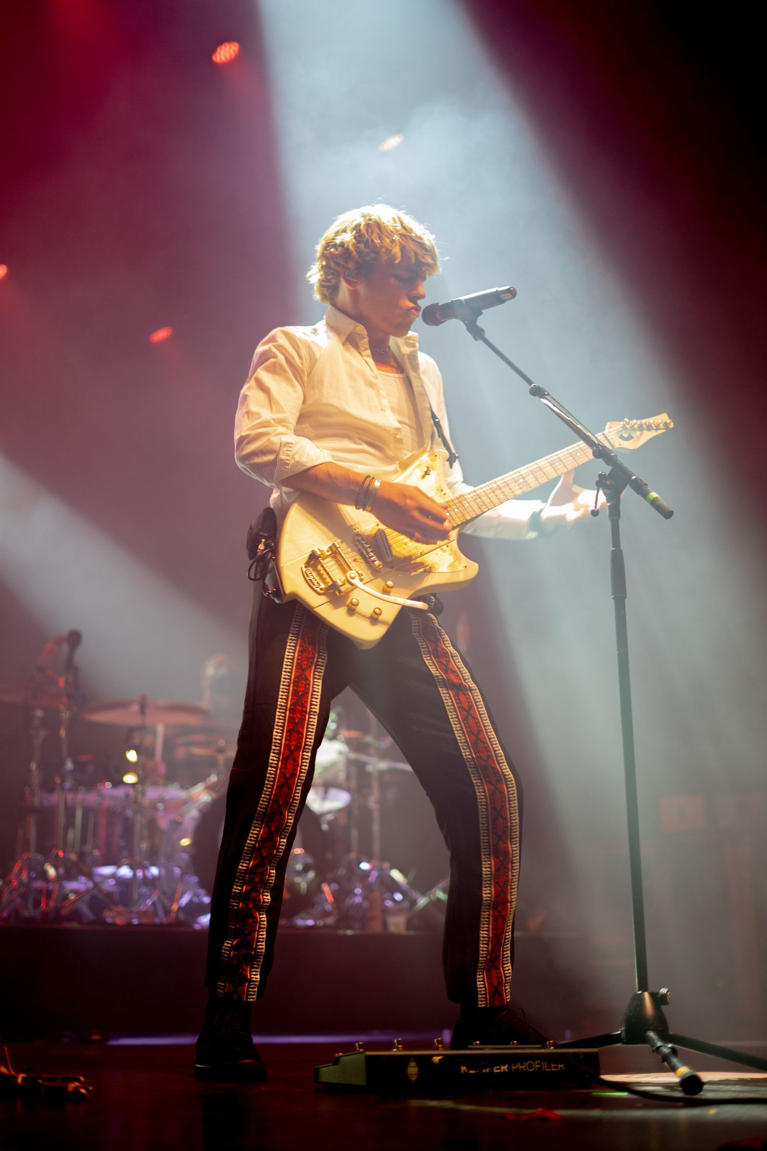 Guitarist in white shirt and striped pants performing under dramatic spotlights on a concert stage.