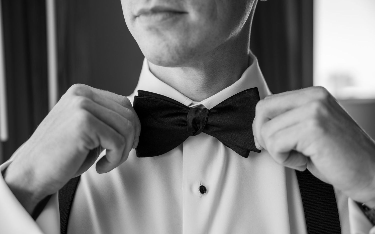 Man in white dress shirt adjusting black bow tie, black and white photo, formal wedding attire.