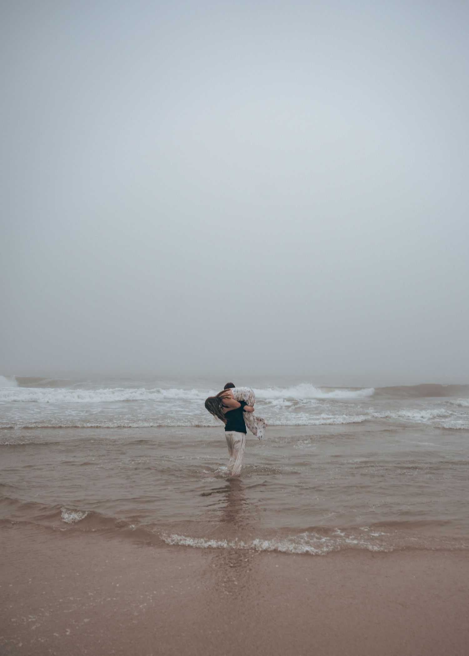 A person carrying someone through misty ocean waves on a foggy overcast beach day.