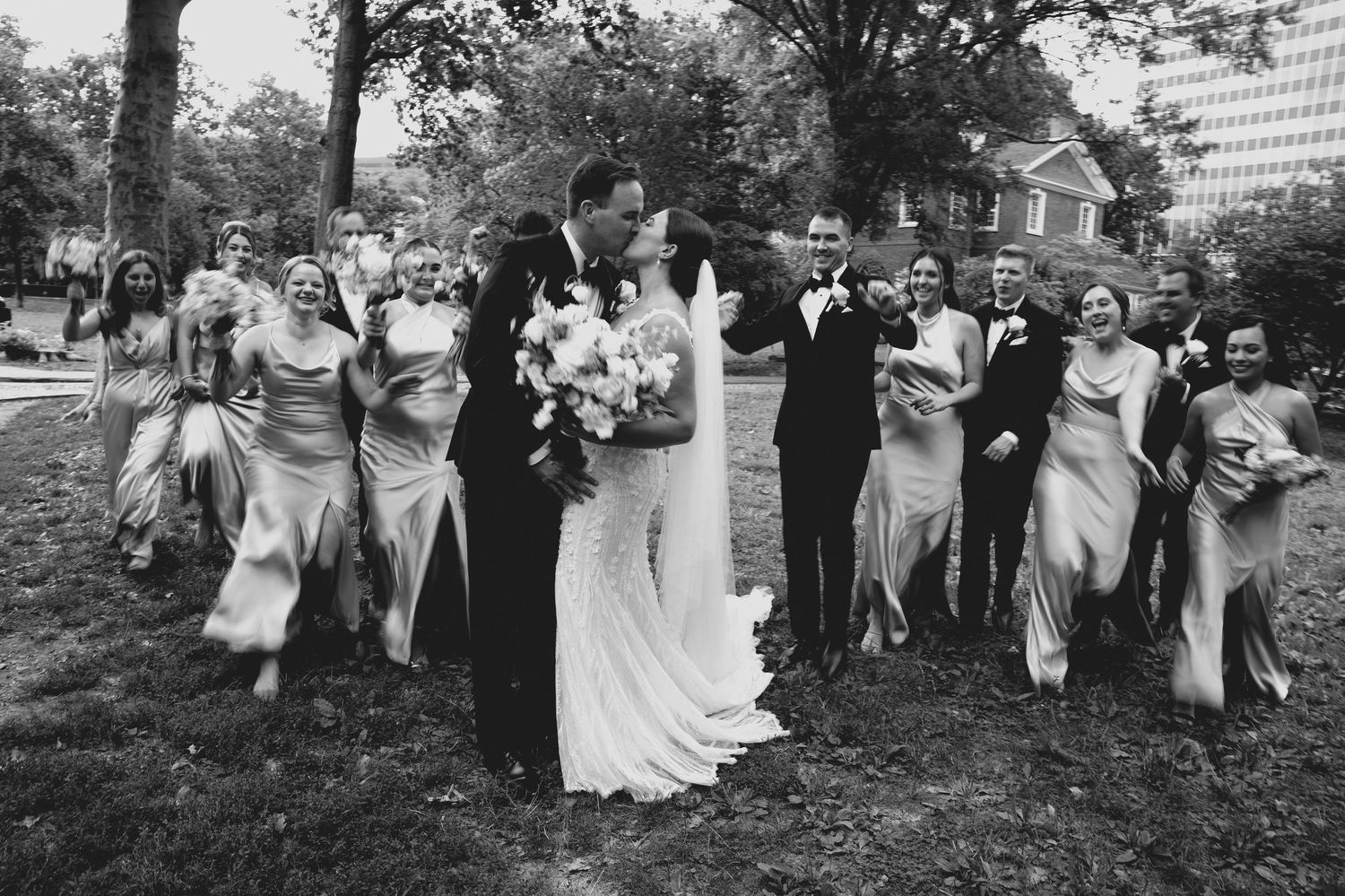 Black and white photo of bride and groom kissing with wedding party outdoors.