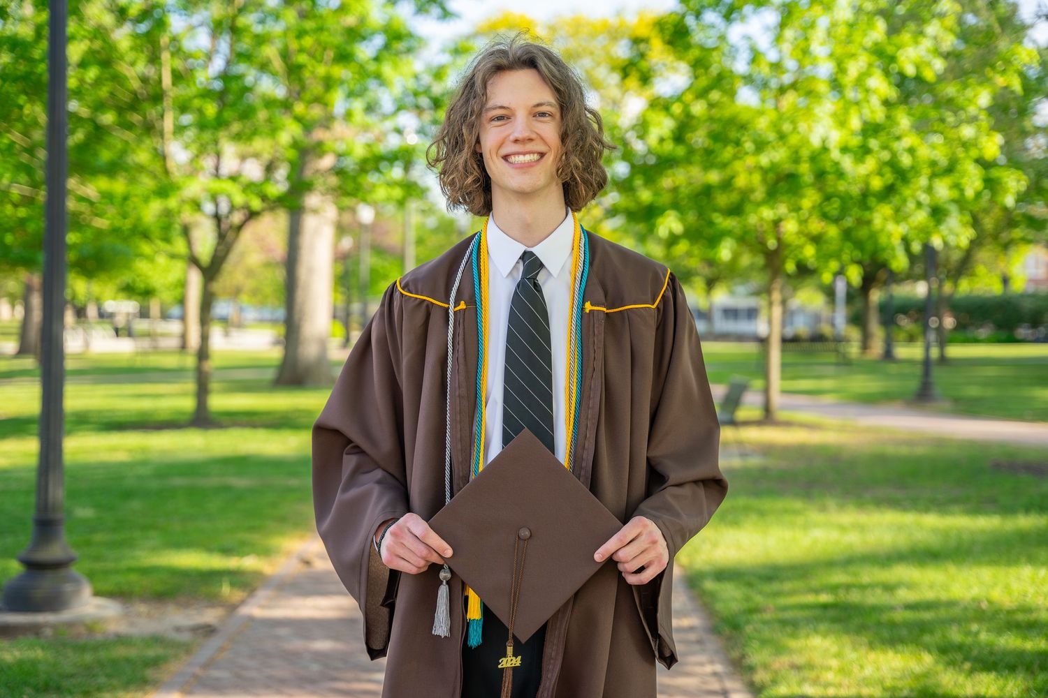 Happy graduate in brown regalia holds mortarboard on a tree-lined campus walkway.