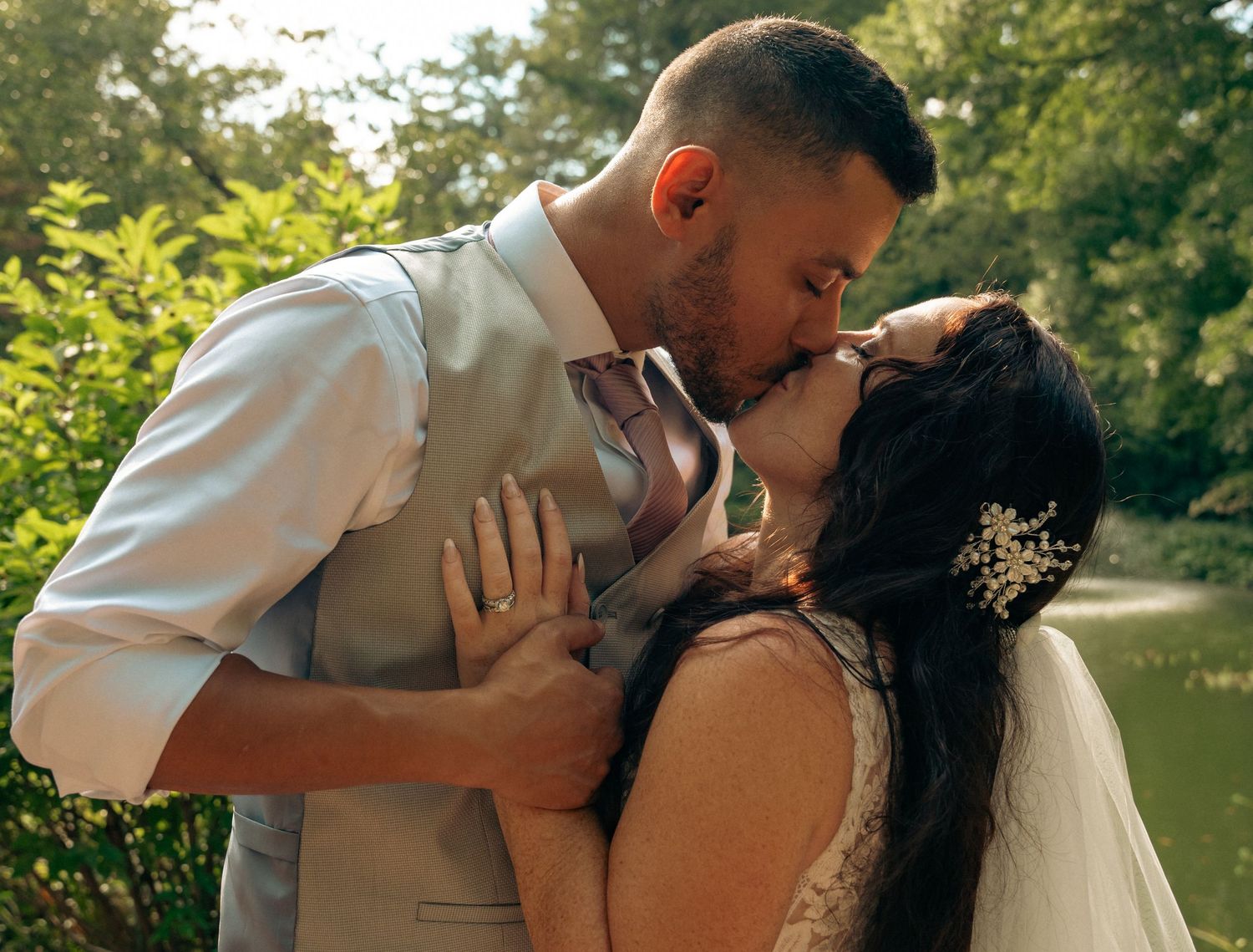 Newlyweds share passionate first kiss surrounded by lush greenery near tranquil garden pond.