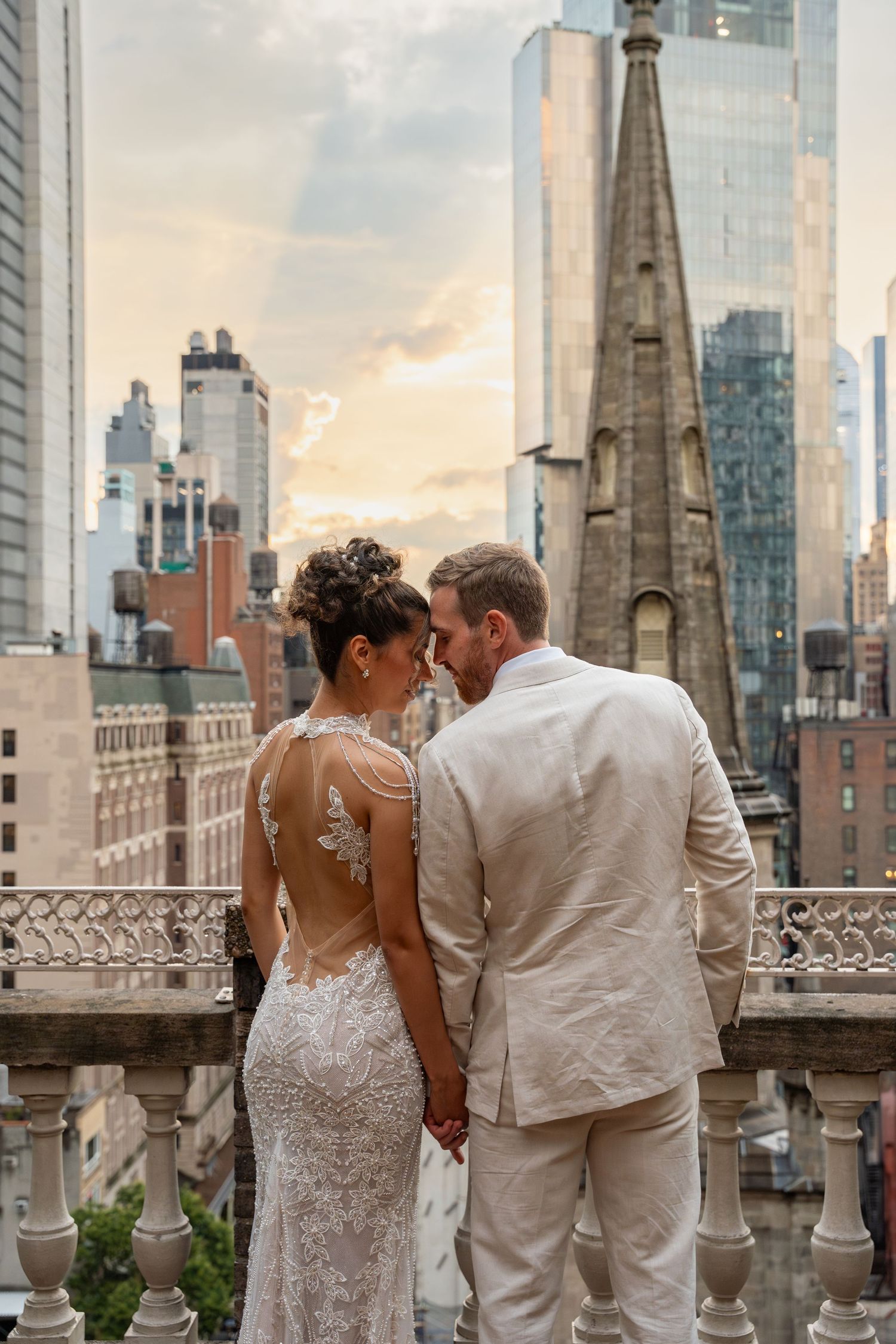 Bride and groom hold hands on a rooftop balcony with NYC skyline at golden hour.