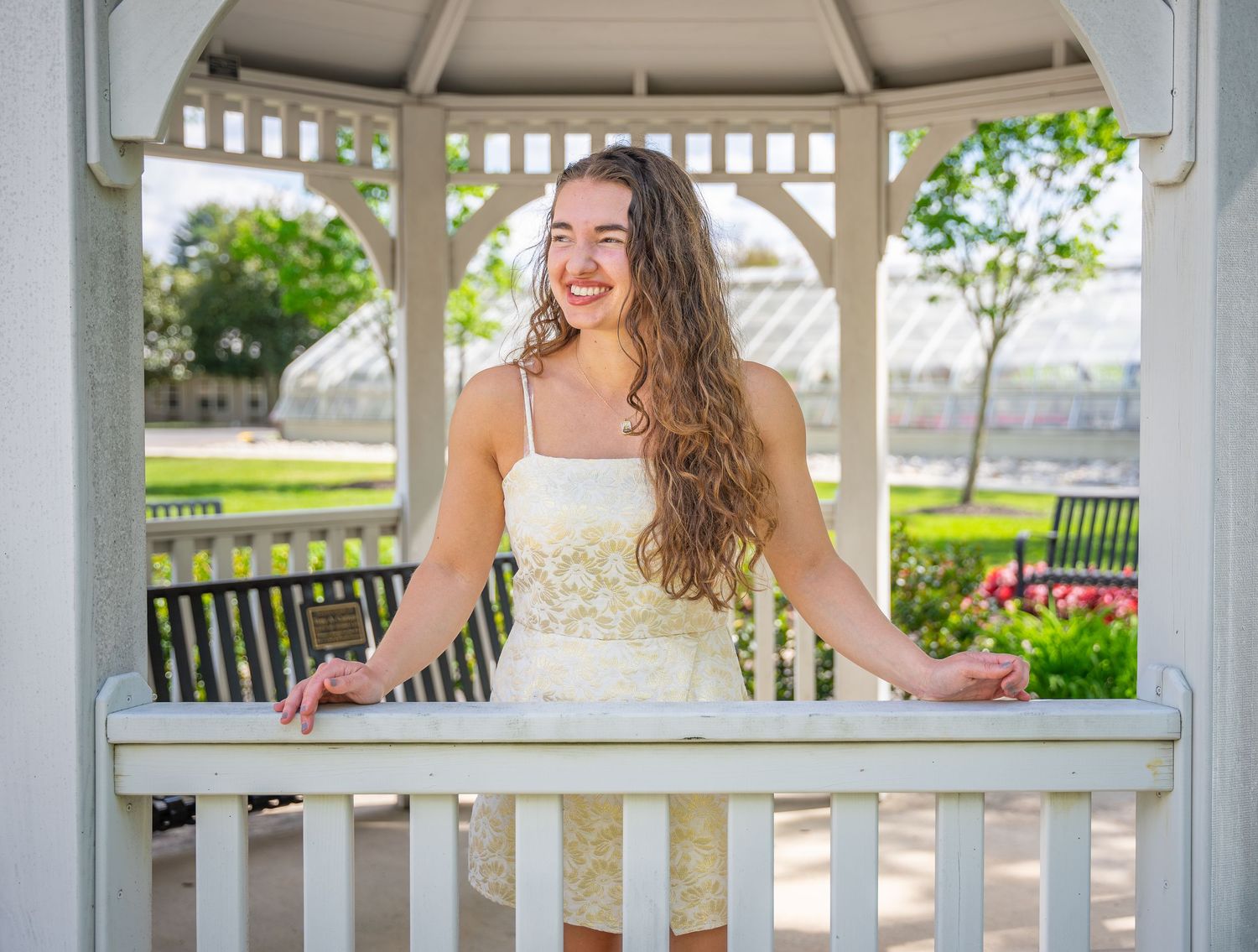 Smiling young woman in white dress posing in a white gazebo in a garden.
