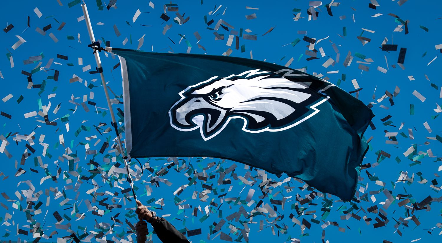 Philadelphia Eagles flag waves proudly against blue sky with silver confetti falling during celebration.