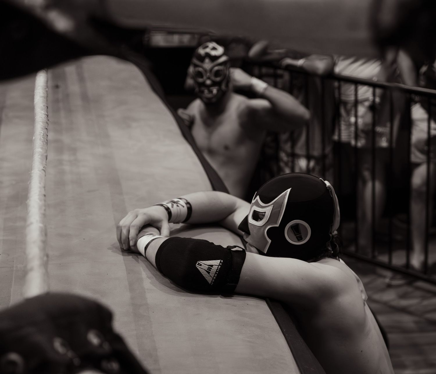 Black and white close-up of masked lucha libre wrestlers resting against ring apron between matches.