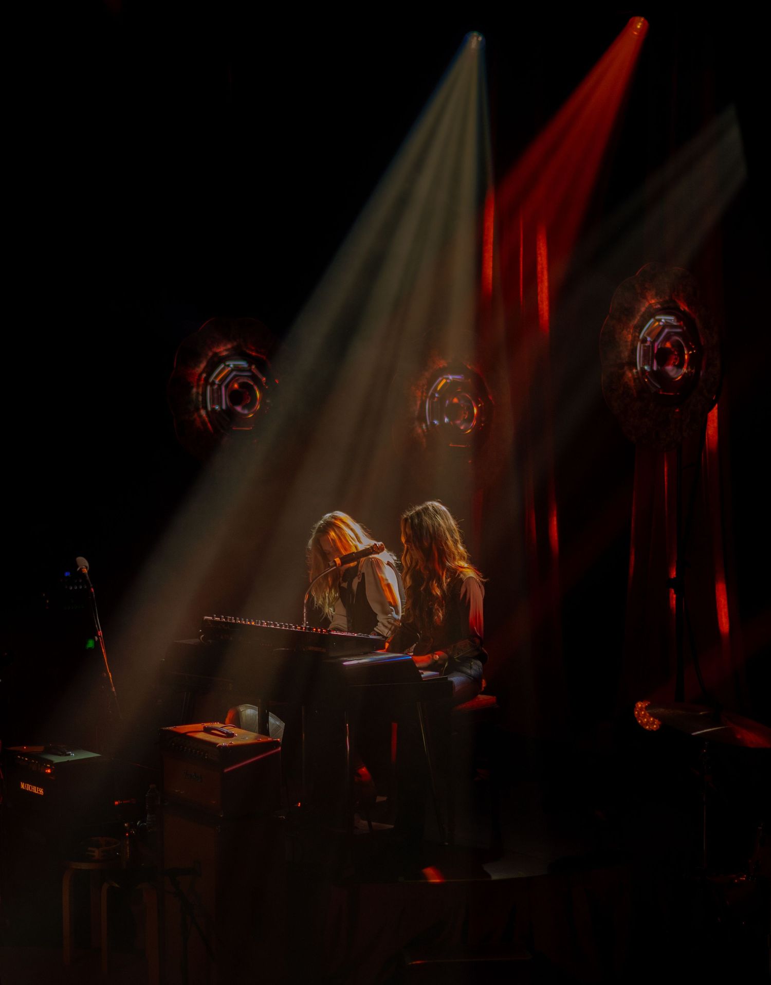 Two musicians share a keyboard on a darkened stage bathed in dramatic red and white spotlights.