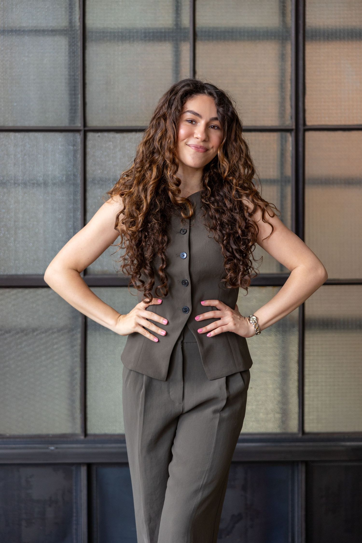 Confident woman with long curly hair in olive green pantsuit posing with hands on hips.
