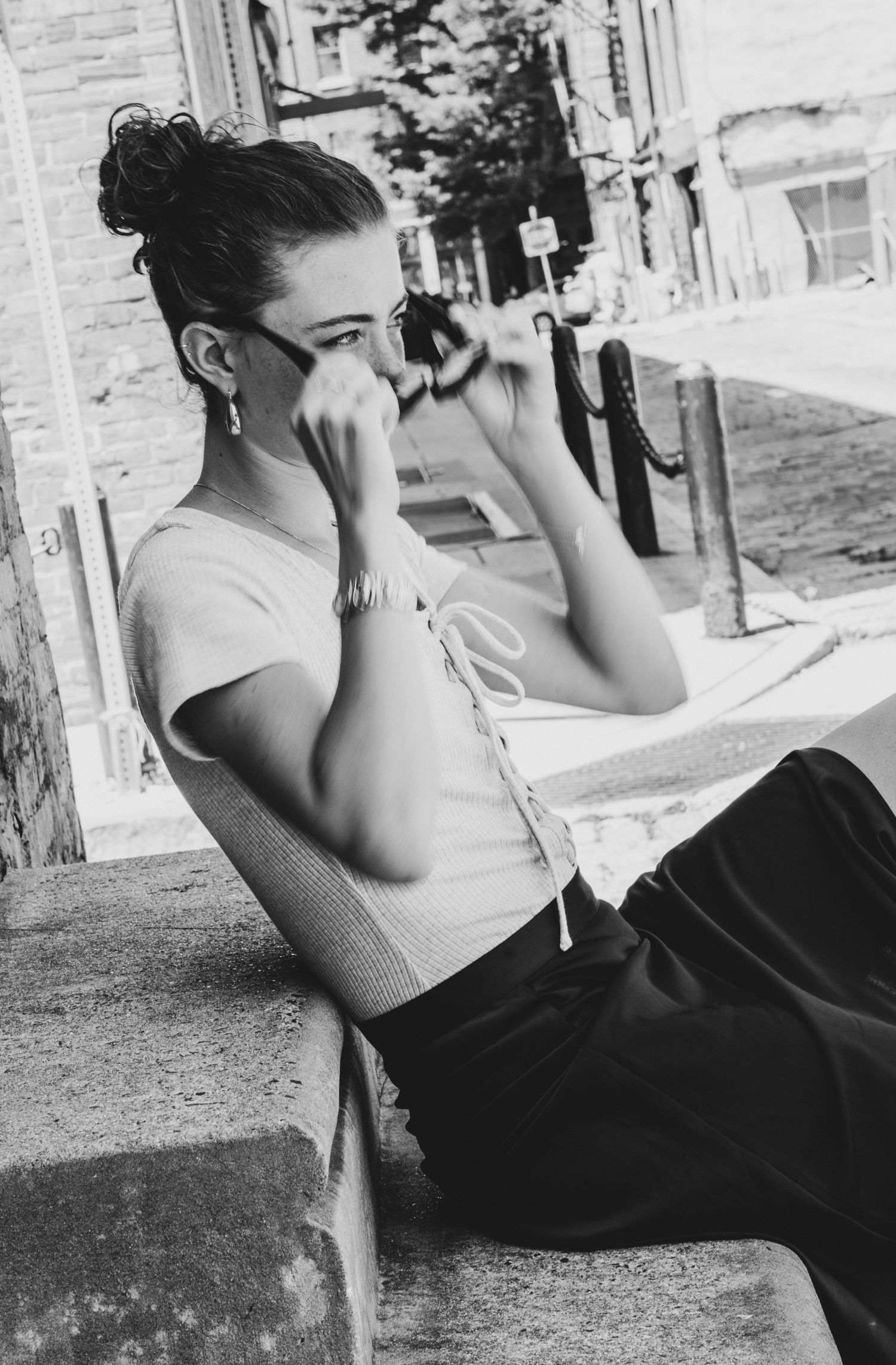 Black and white photo of young woman sitting outdoors adjusting sunglasses on urban steps.
