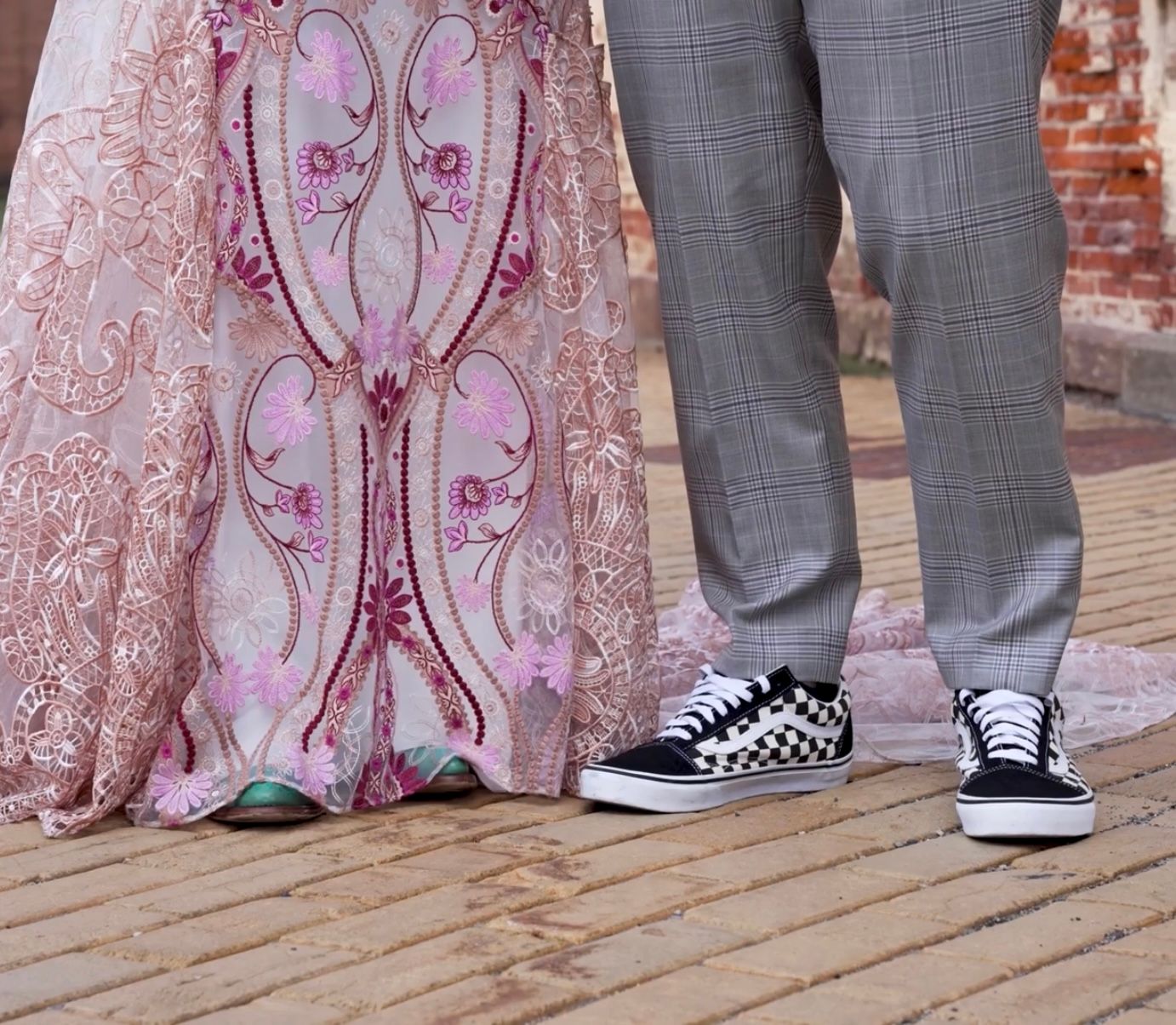 Close-up of bride in pink embroidered gown beside groom wearing checkered Vans sneakers on brick.