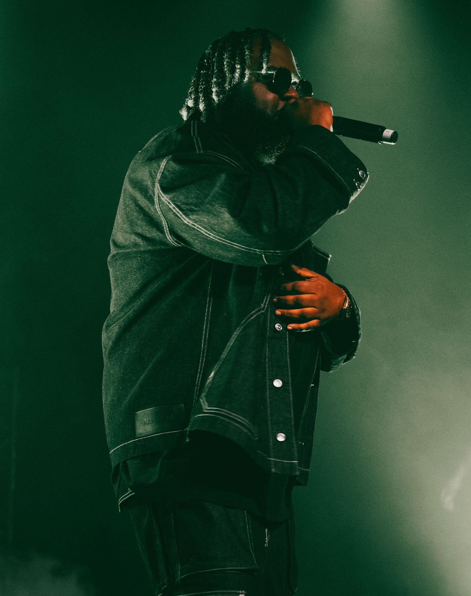 Rapper performing on stage wearing a dark denim jacket and sunglasses under green stage lighting.