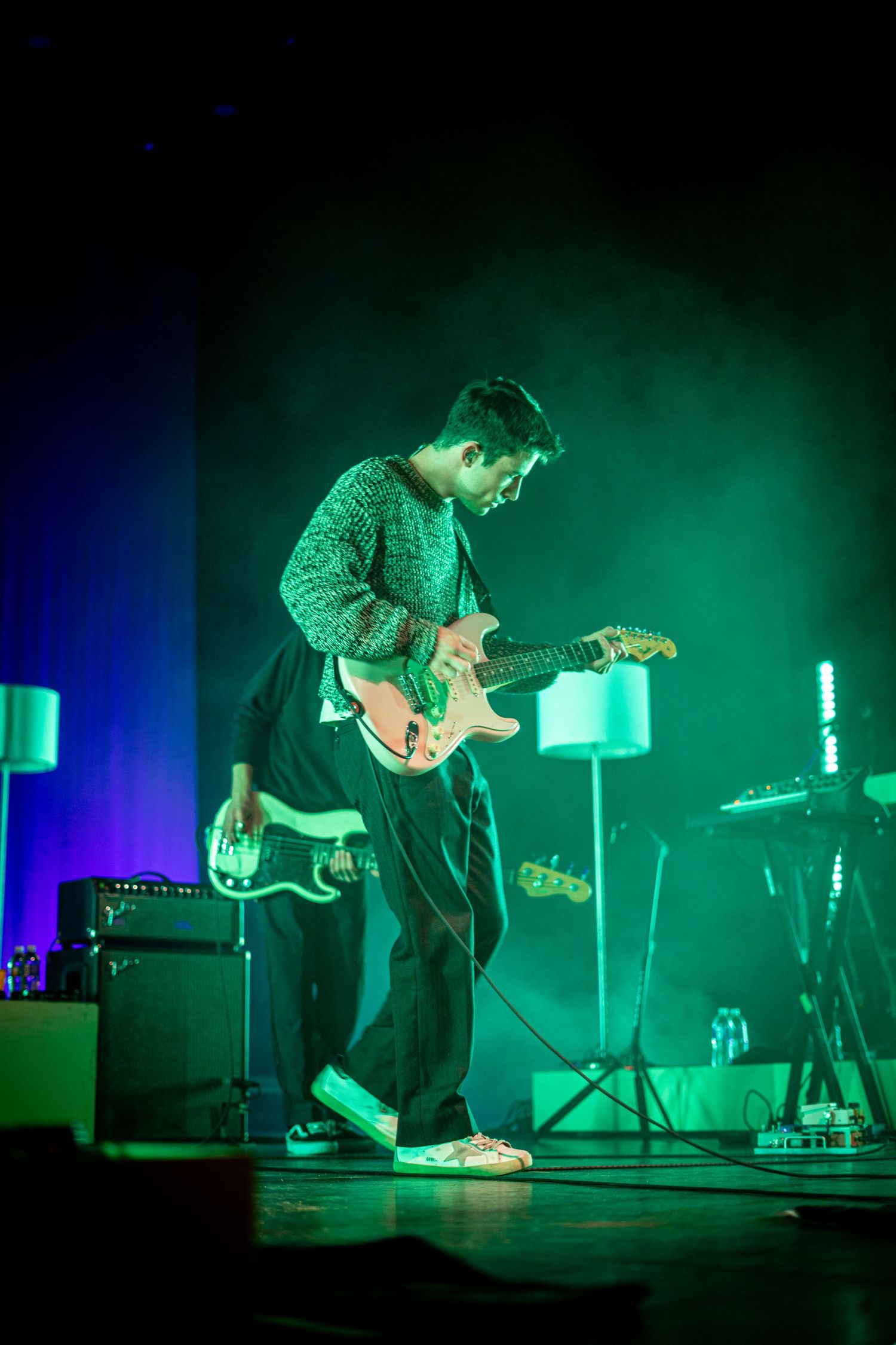 Guitarist playing electric guitar on stage surrounded by green and blue atmospheric stage lighting.