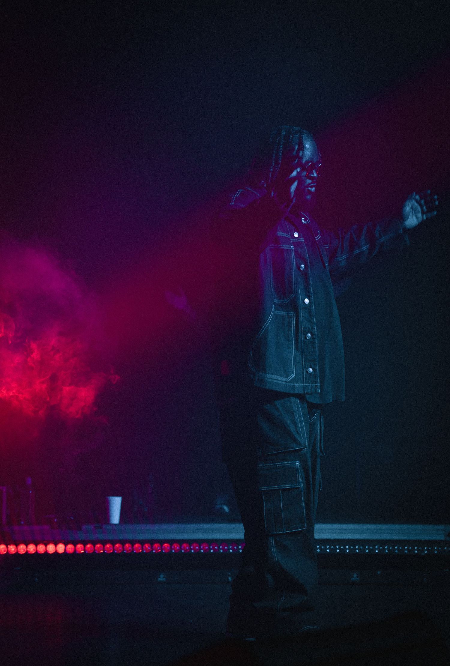 Performer with dreadlocks standing on stage with arms outstretched in vivid red and purple lighting.