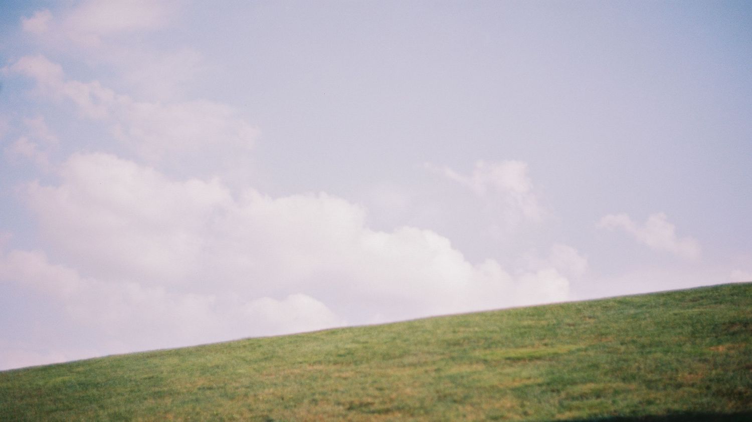 Minimalist landscape of green grassy hill against soft pastel blue sky with white clouds.