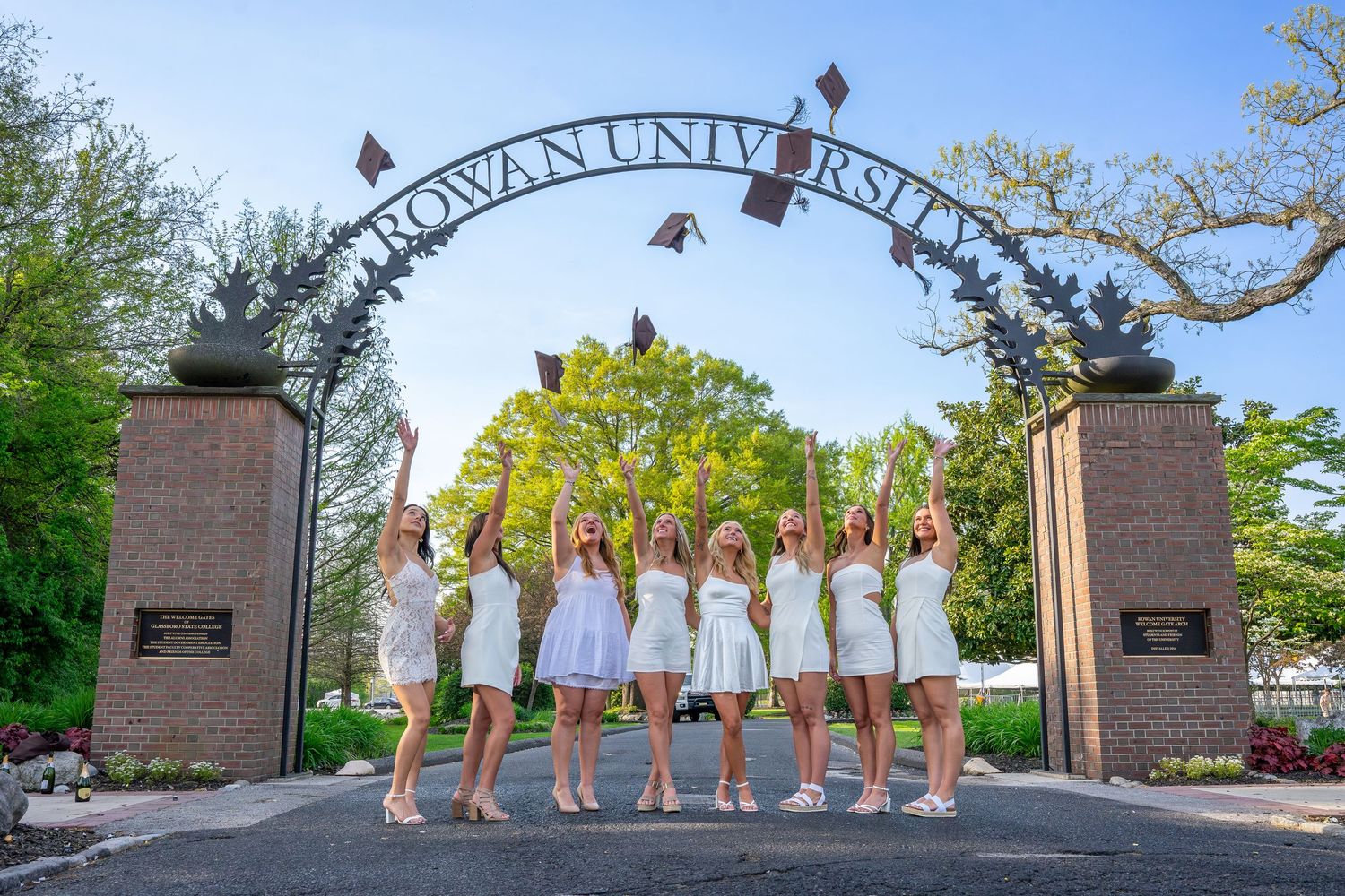 Nine graduates in white dresses tossing caps under Rowan University entrance gate.