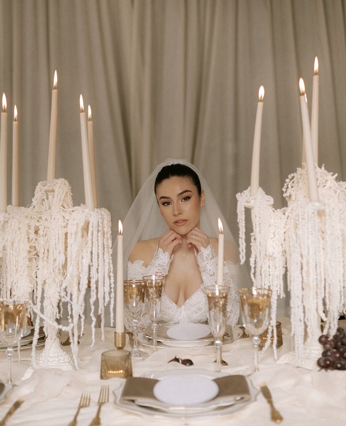 Elegant bride in veil seated at candlelit wedding table with tall white tapers and crystal decor.
