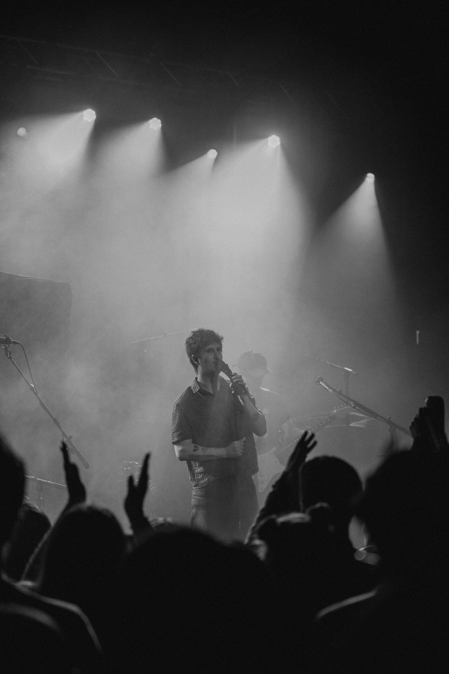 Black and white concert photograph of a guitarist under stage light beams above a crowd with raised hands at a Philadelphia music venue