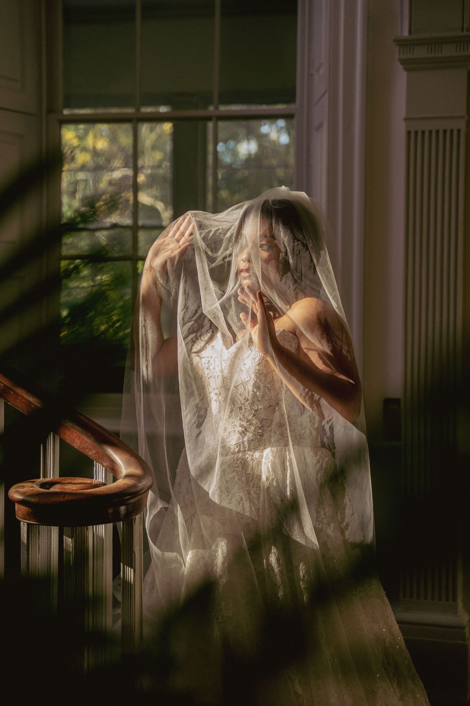 Bride lifts sheer veil in soft golden light by staircase window in moody, intimate indoor portrait.