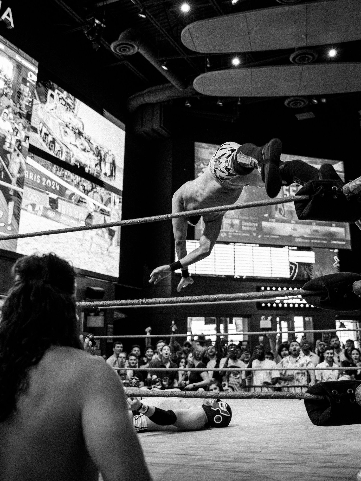 Black and white photo of masked lucha libre wrestler performing aerial move over opponent in wrestling ring.