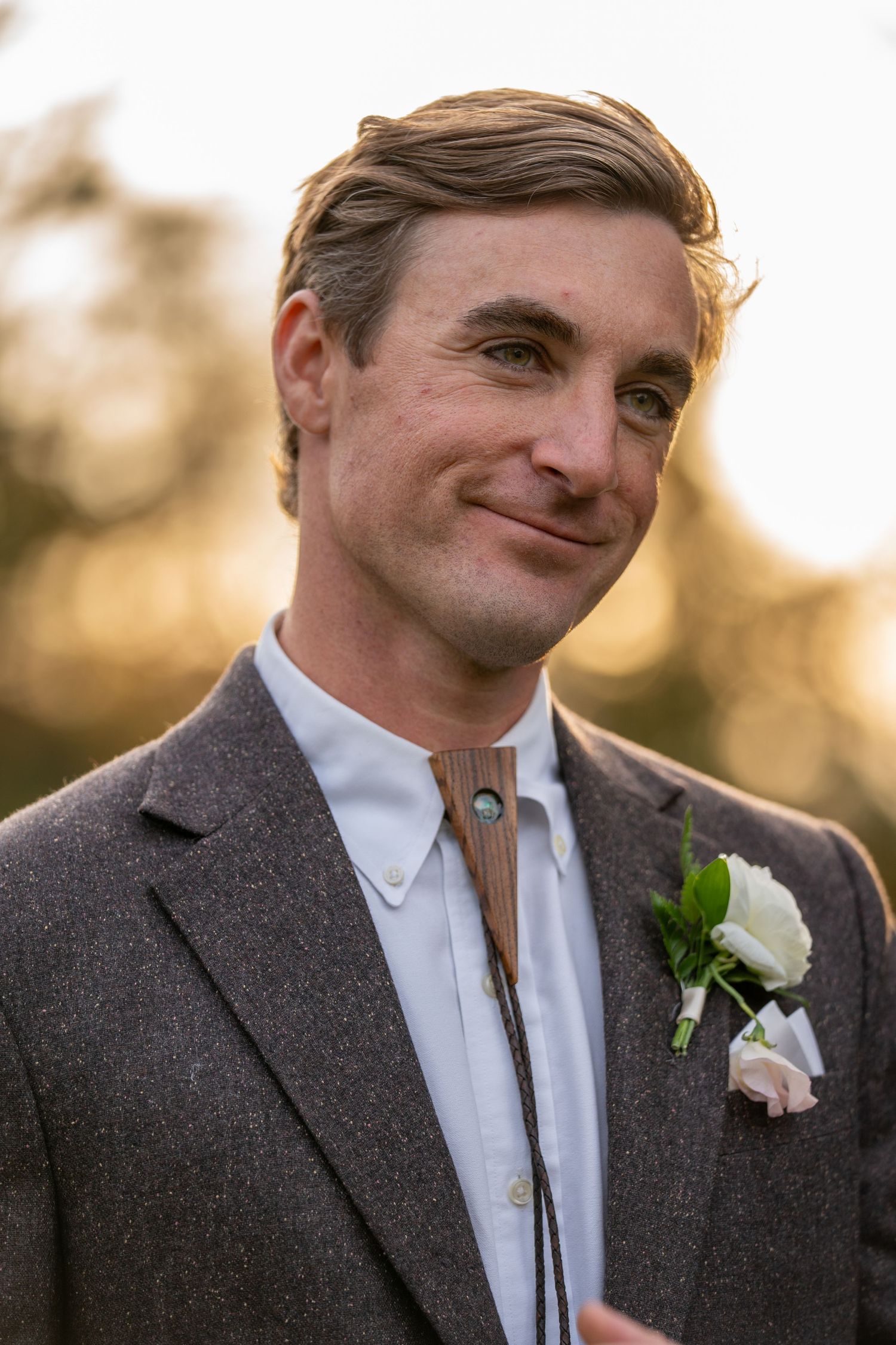 A dapper groom in a grey tweed suit with bolo tie and white rose boutonniere smiles at golden hour.