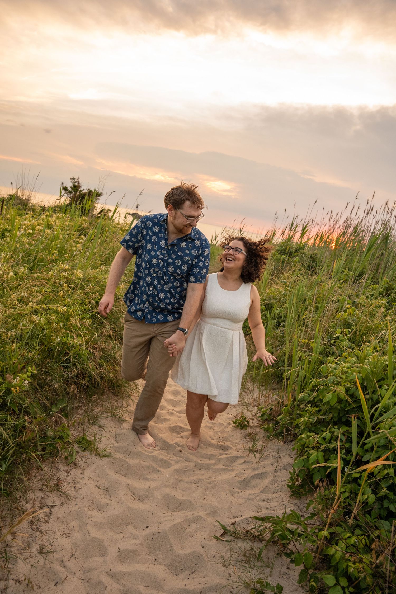 Couple holding hands and laughing while walking on sandy beach path at sunset with lush green grass.