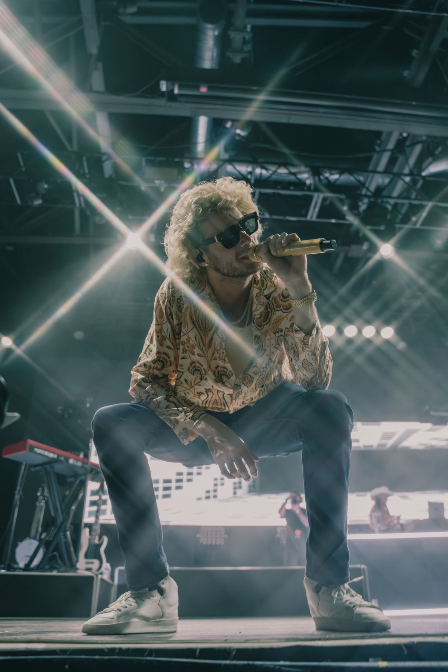 Singer performs on stage wearing patterned shirt and sunglasses under bright concert lights.