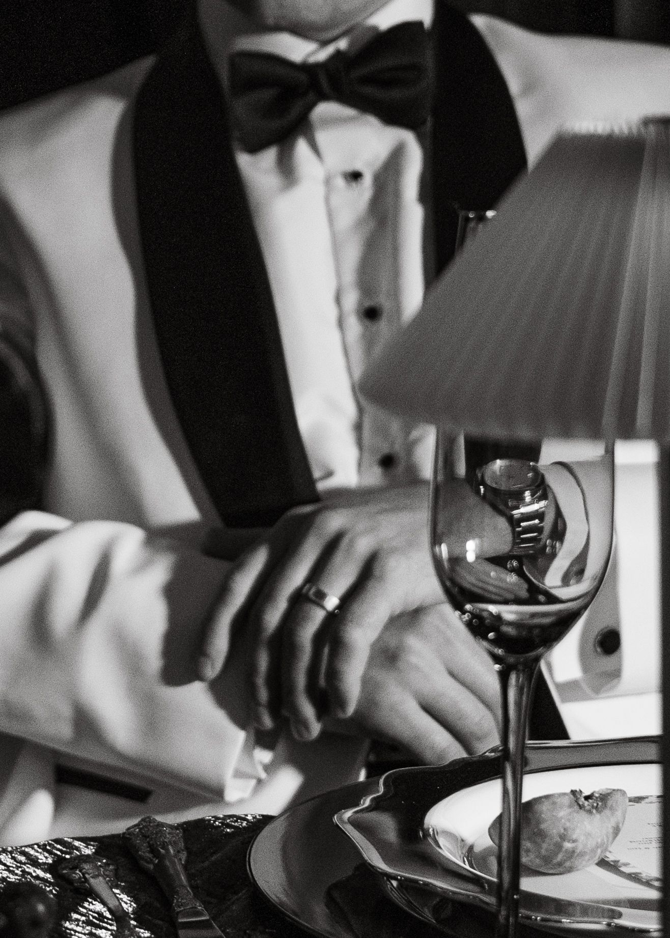 Black and white photo of a groom in a tuxedo with bow tie reaching for a wine glass at a table.