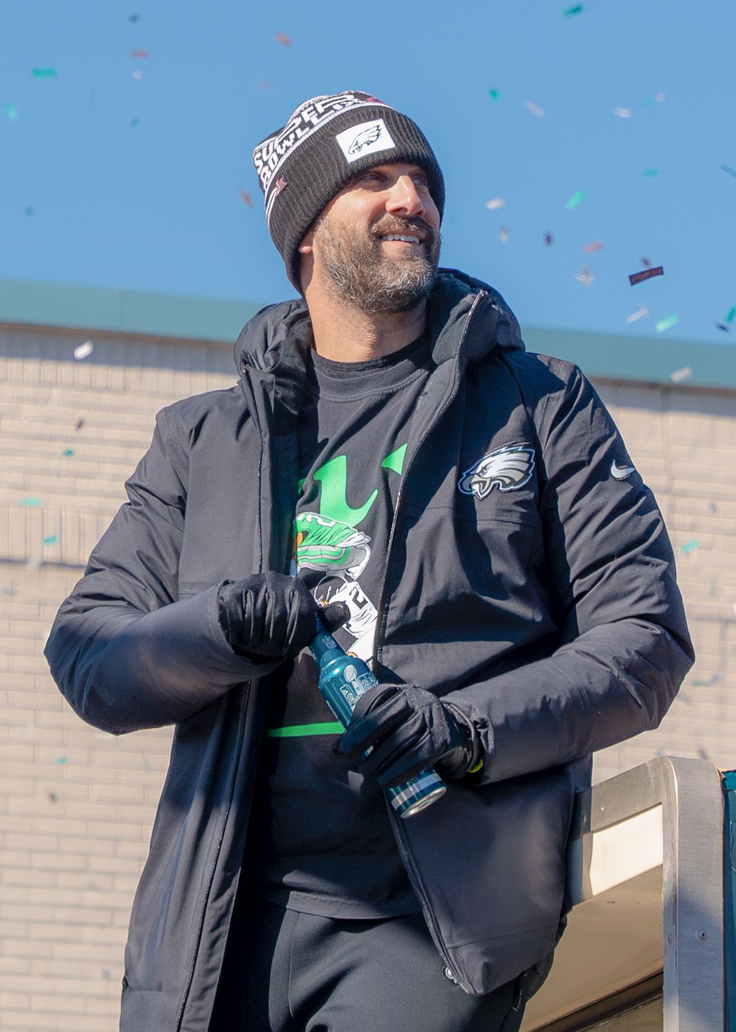 Bearded man in Philadelphia Eagles gear smiles during confetti-filled outdoor championship parade celebration.
