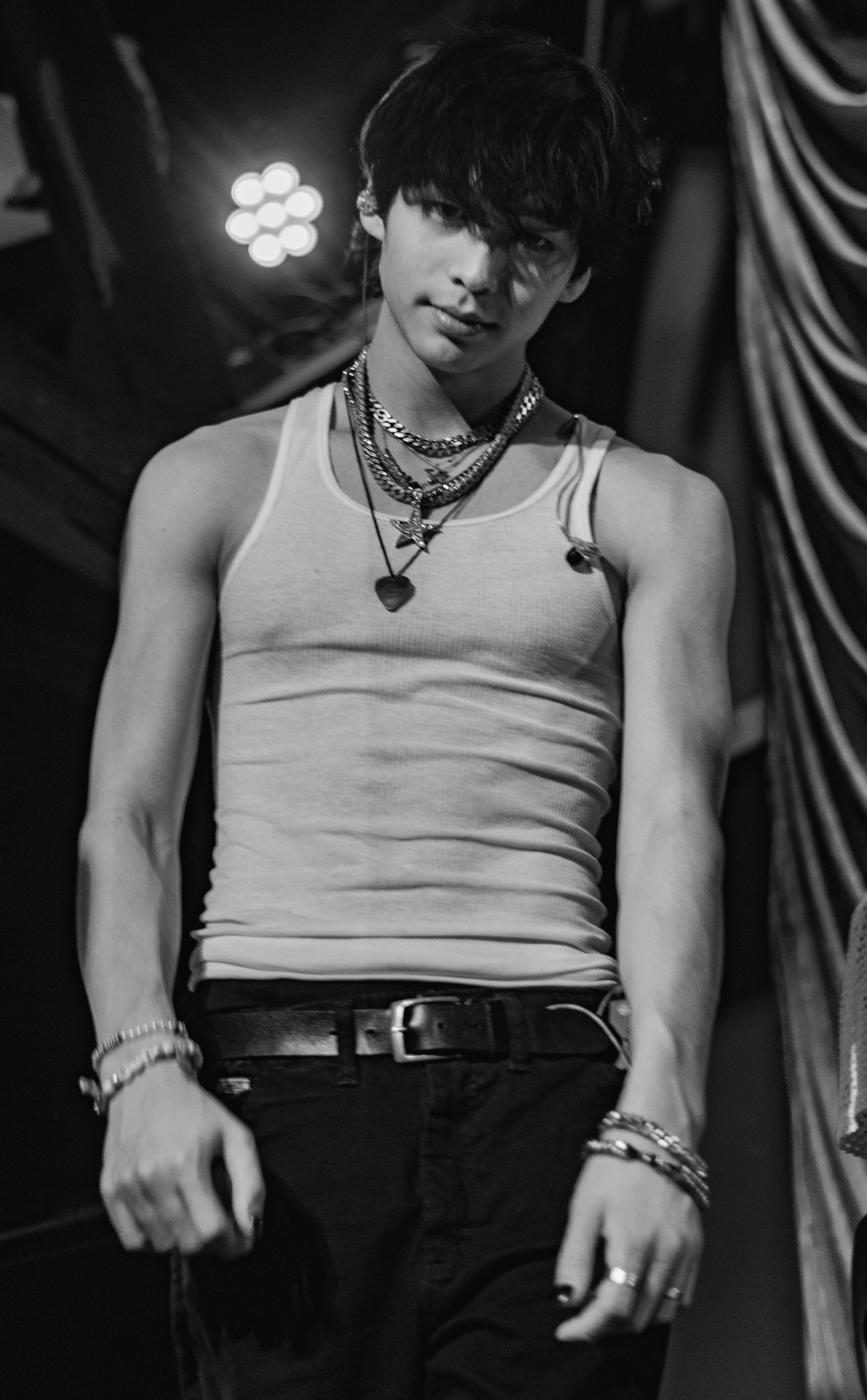 Black and white photo of a young male performer in a tank top and chains on stage under dramatic lighting.
