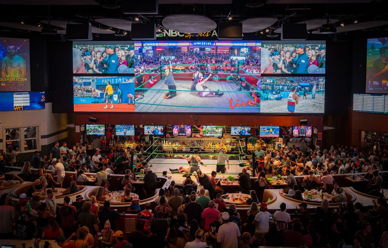 Large sports bar crowd watches lucha libre wrestling match under massive LED screens at NBA arena venue.