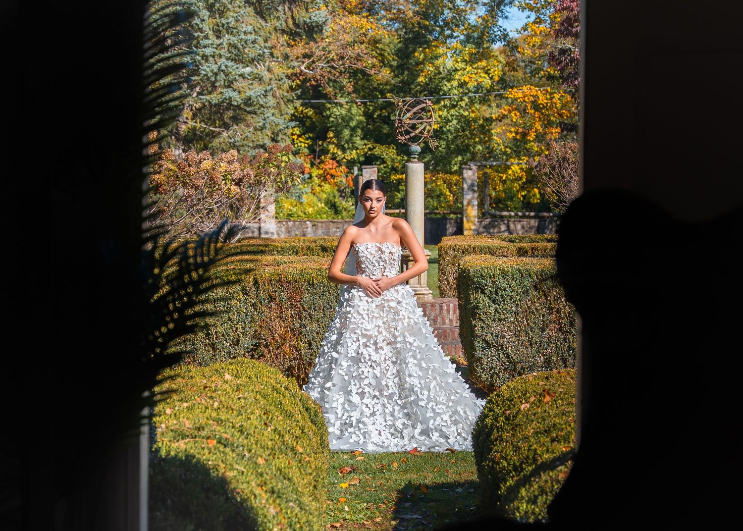 Woman in white floral ball gown poses in lush garden with autumn foliage and trimmed hedges.