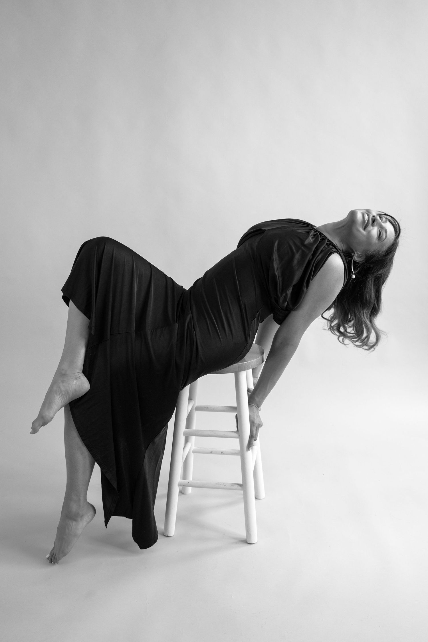 Black and white photo of a woman in a black dress dramatically arching back on a white stool in a studio.