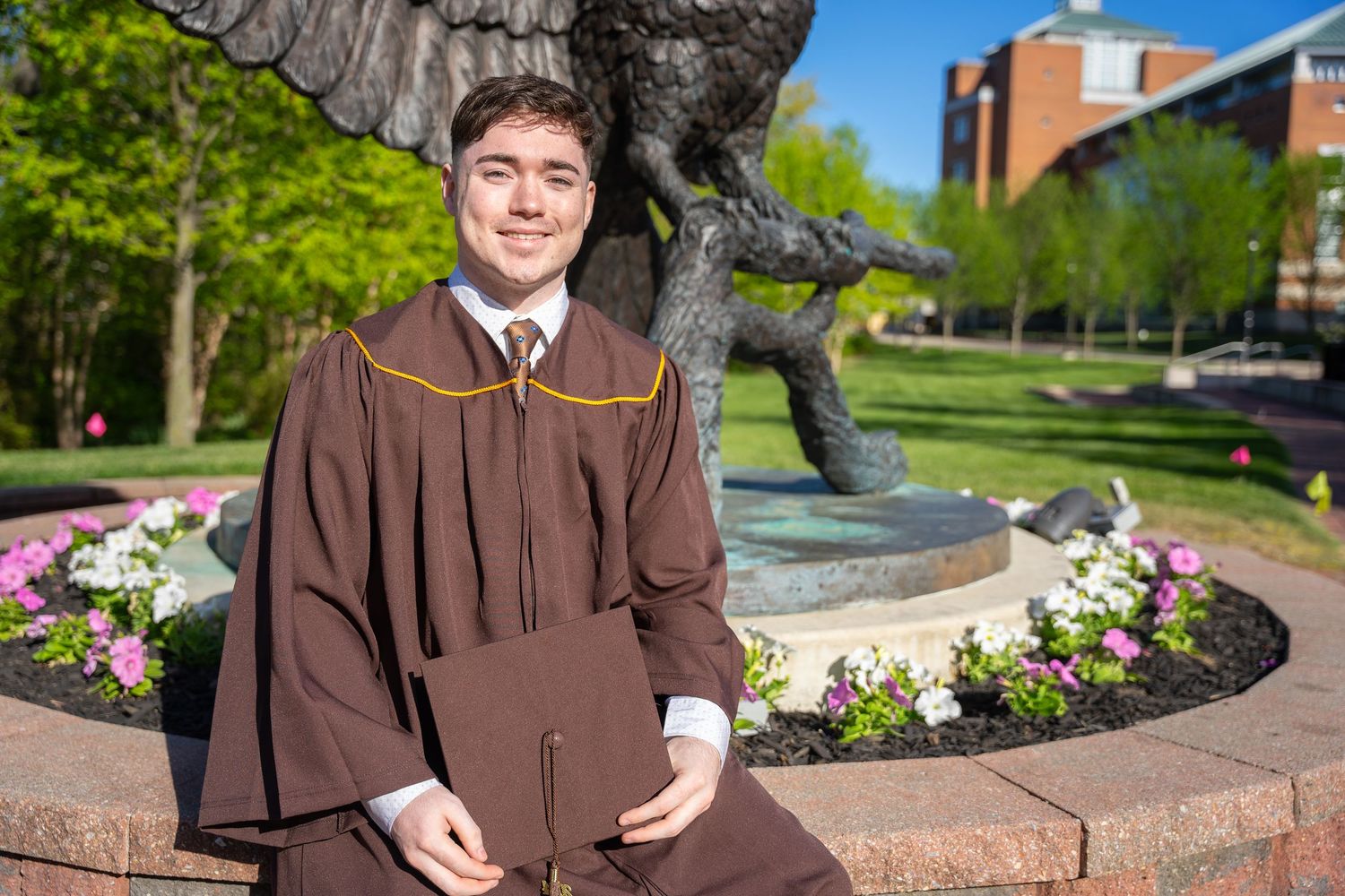 Proud graduate in brown cap and gown sits by campus statue on sunny day.