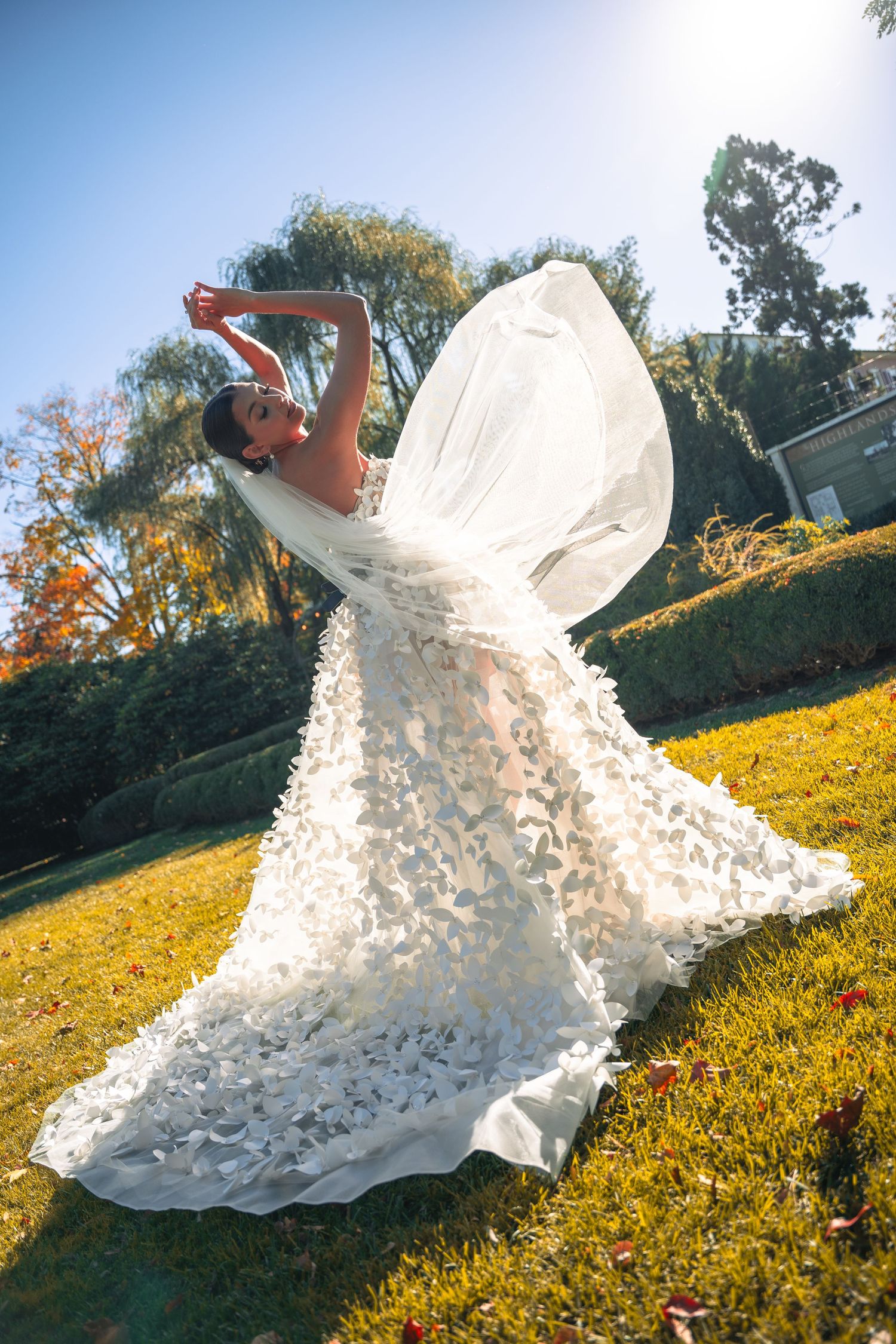 Bride twirls floral applique wedding gown with flowing veil in sunny outdoor garden setting.
