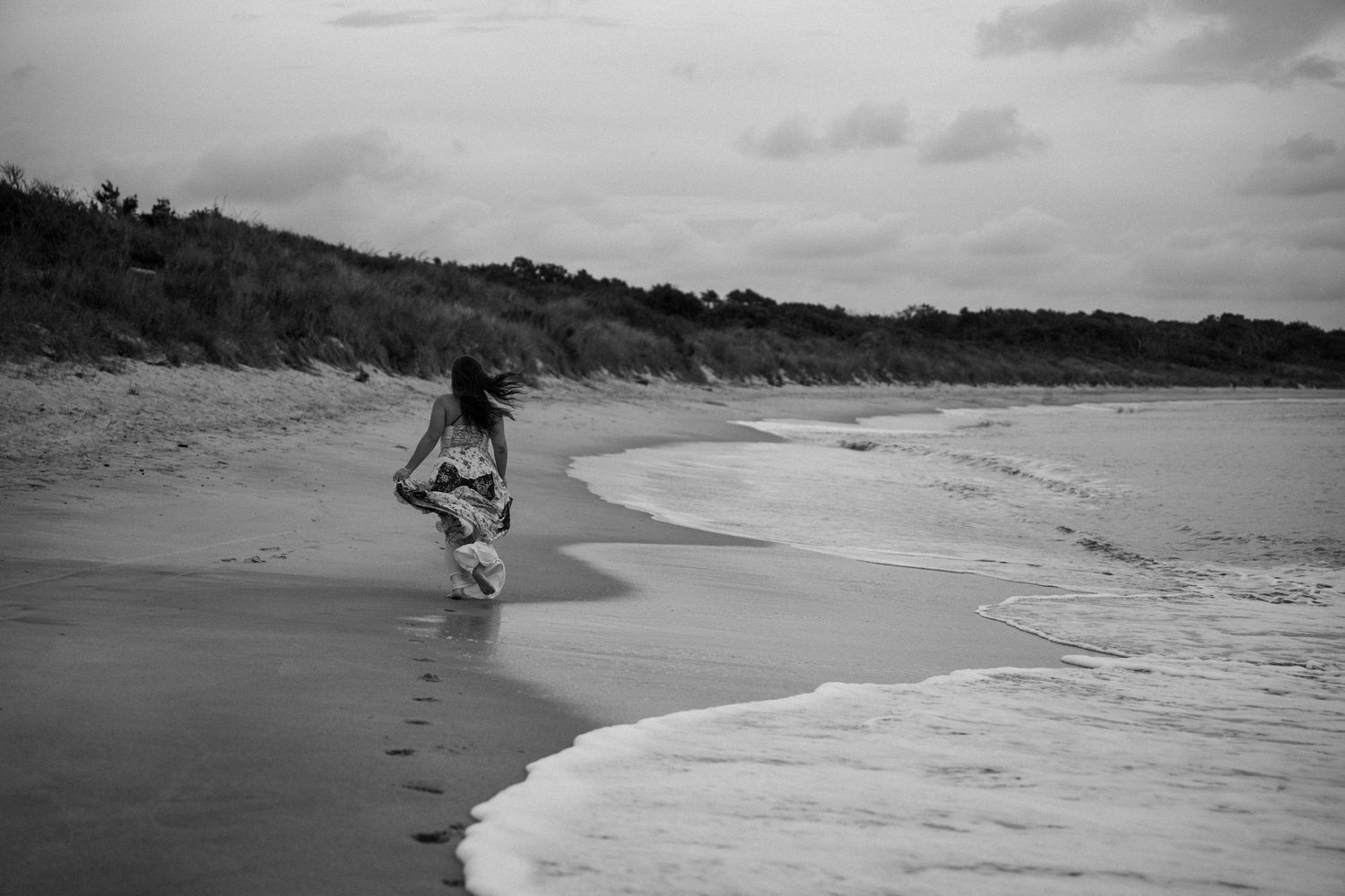 Black and white photo of a woman walking alone along a sandy beach shoreline.