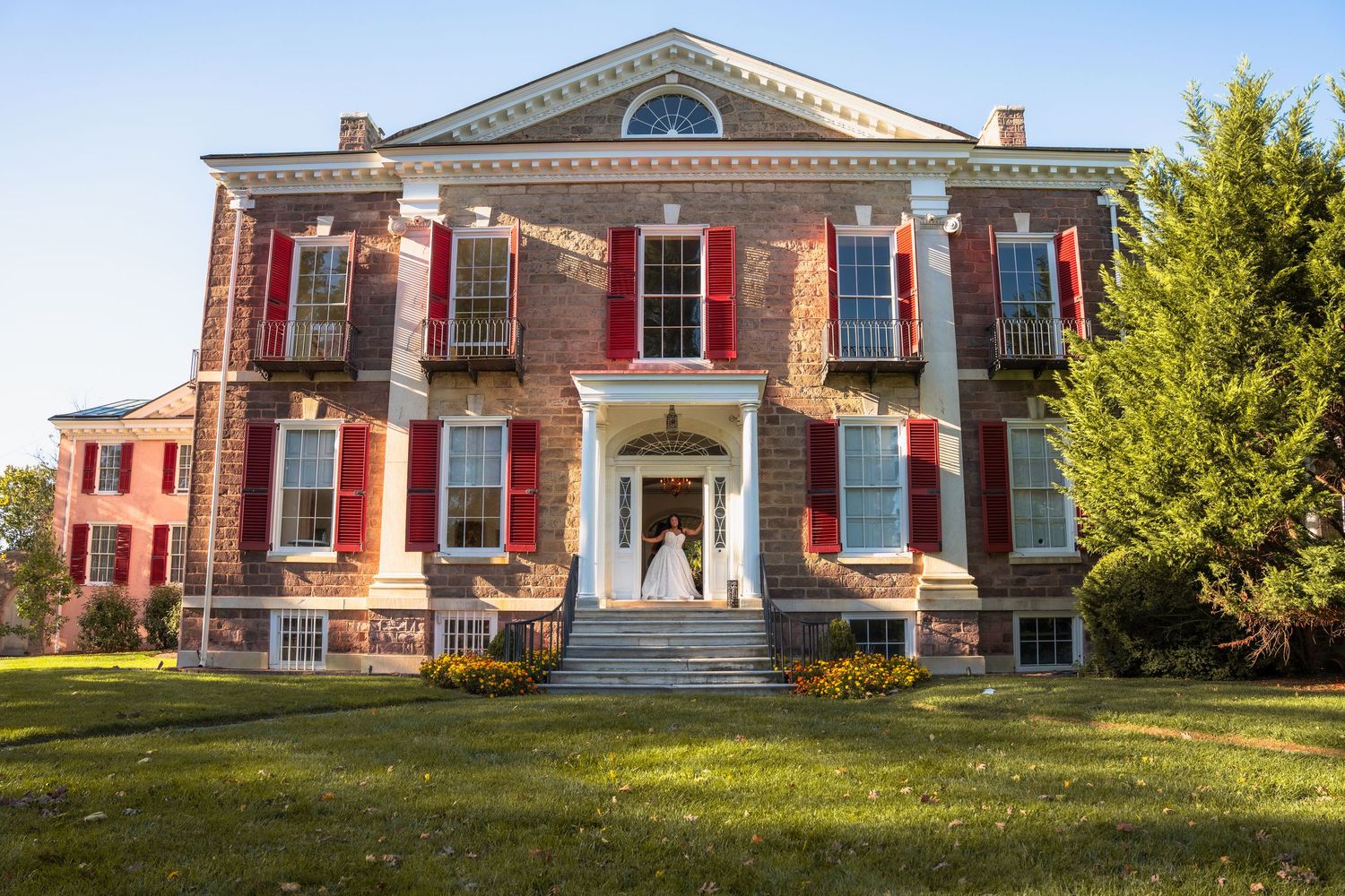 Bride stands in the doorway of a grand red-brick colonial mansion with red shutters on a sunny day.