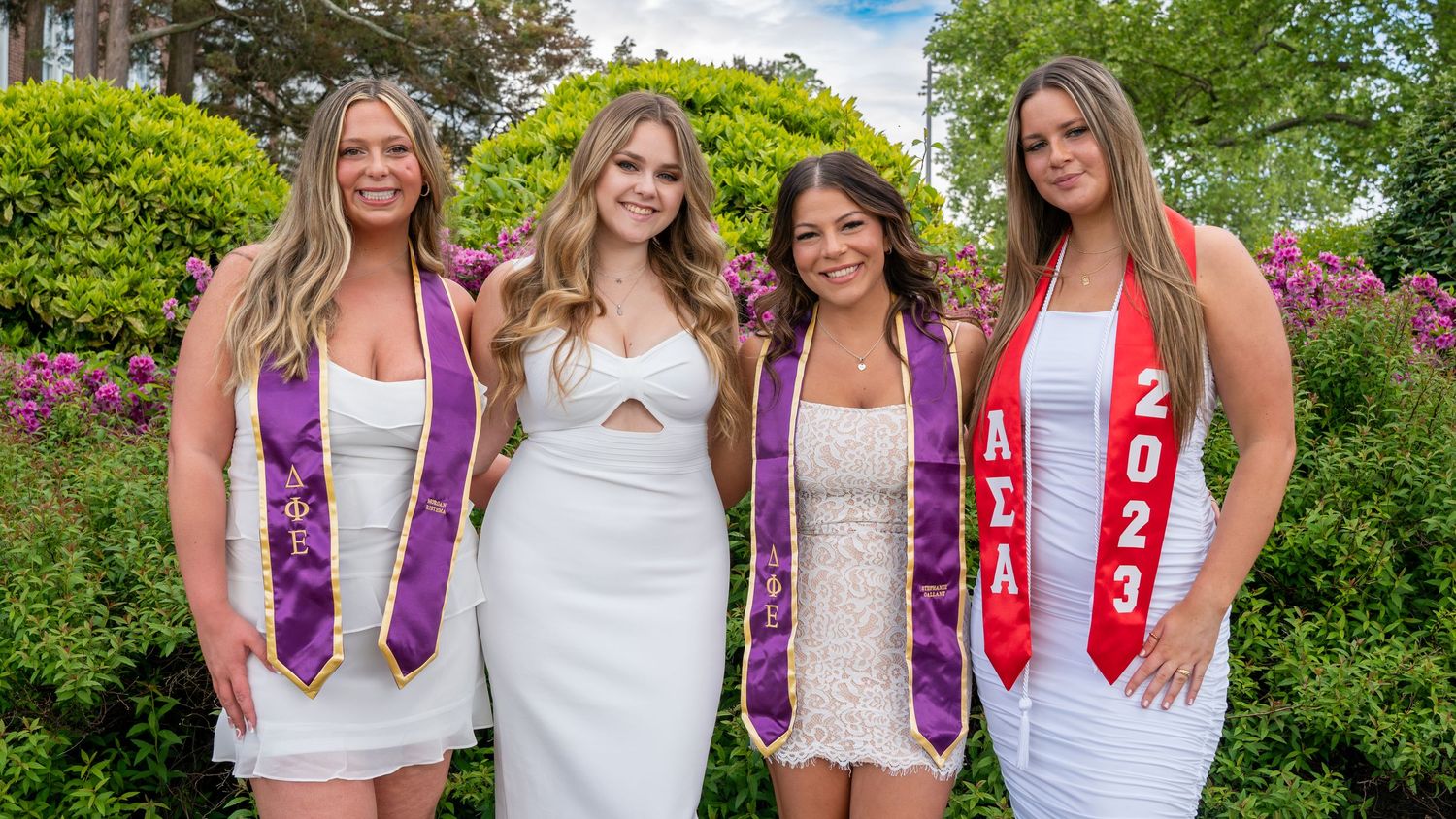 Four smiling women in white dresses wearing graduation stoles posing outdoors at a ceremony.