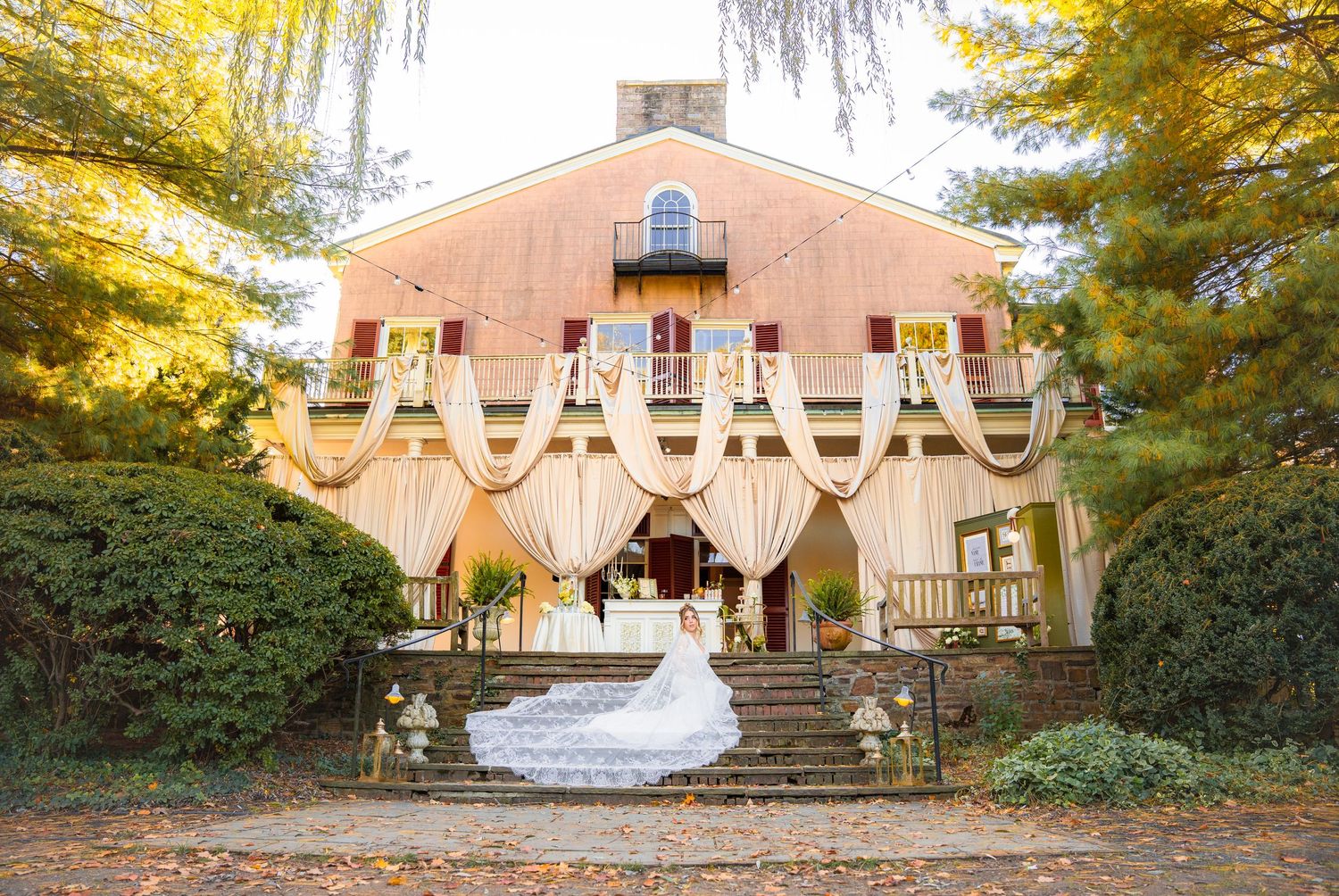 Bride in flowing gown with cathedral veil poses before a draped historic venue in autumn.