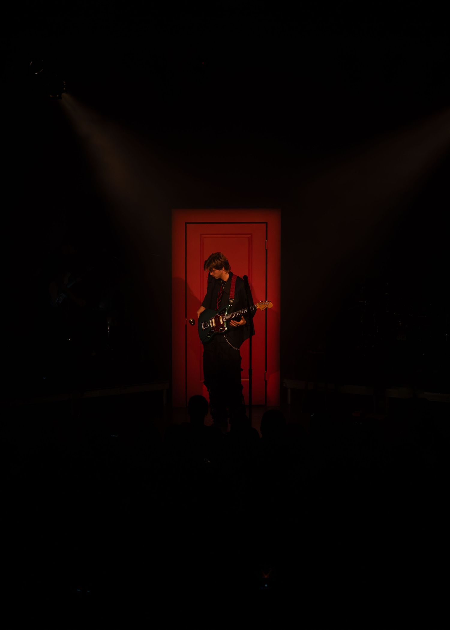 Guitarist performing on dark stage illuminated by dramatic red doorway and spotlight beams.