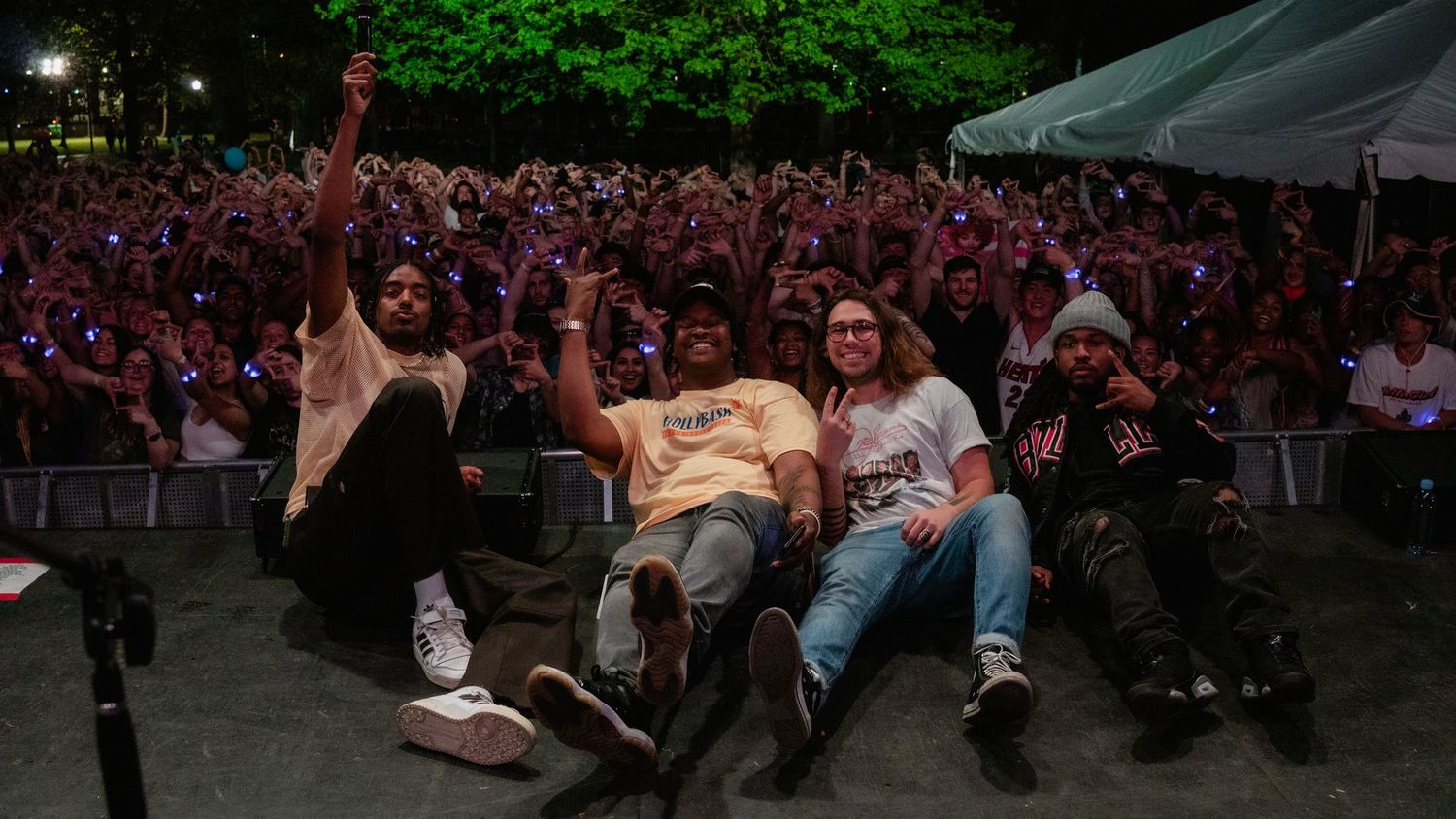 Four performers sit at the edge of a stage in front of a large enthusiastic concert crowd at night.