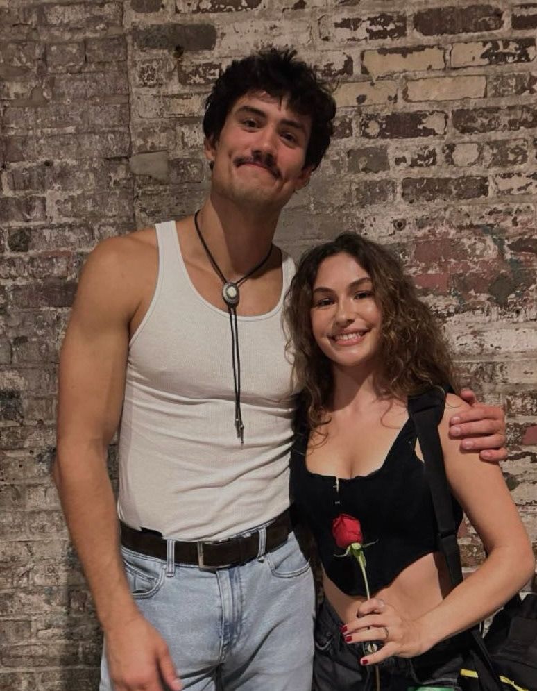 Young couple posing together against brick wall, man holding woman with rose.