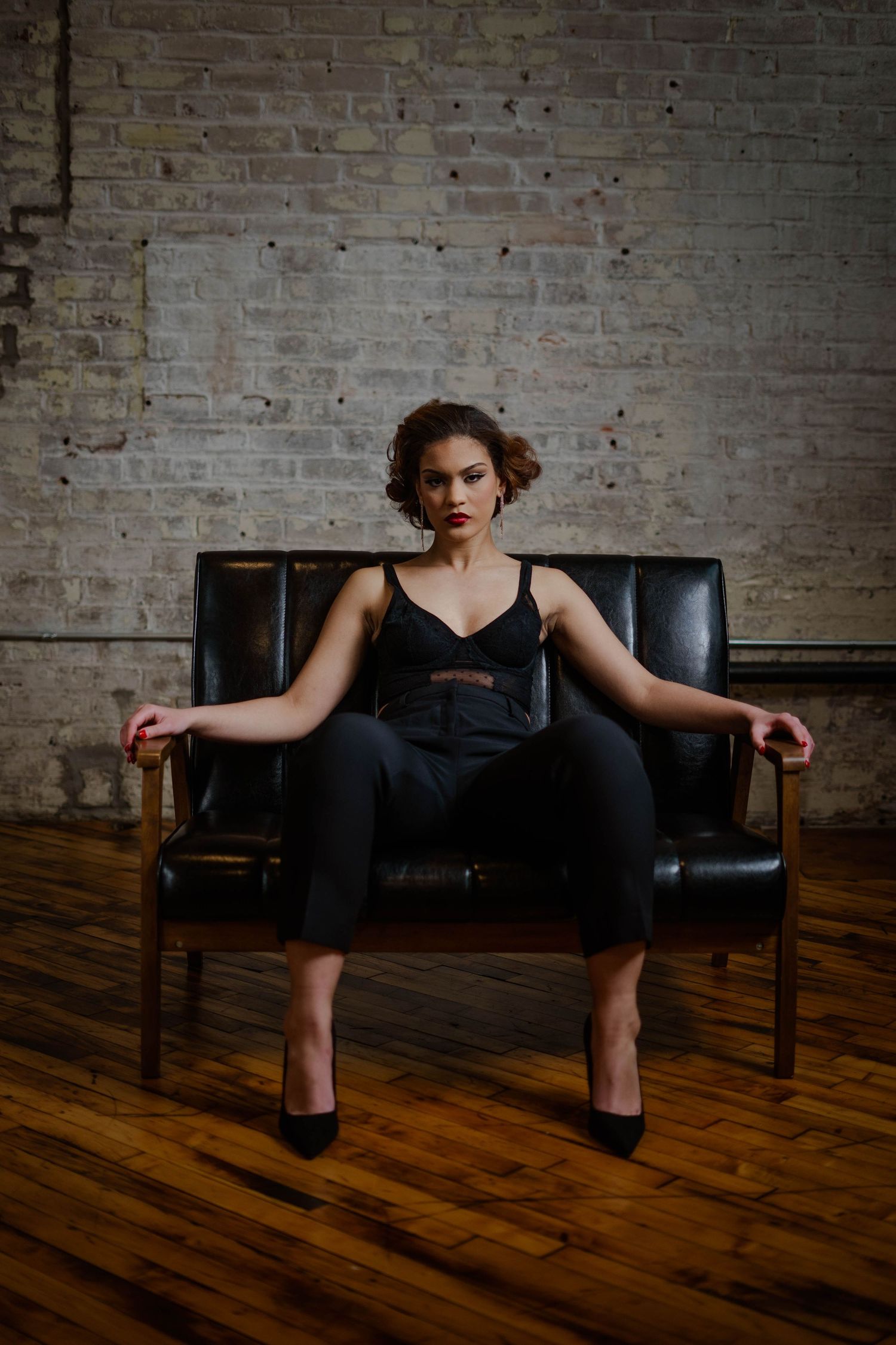 Confident woman in black outfit seated in leather chair against rustic brick loft wall.