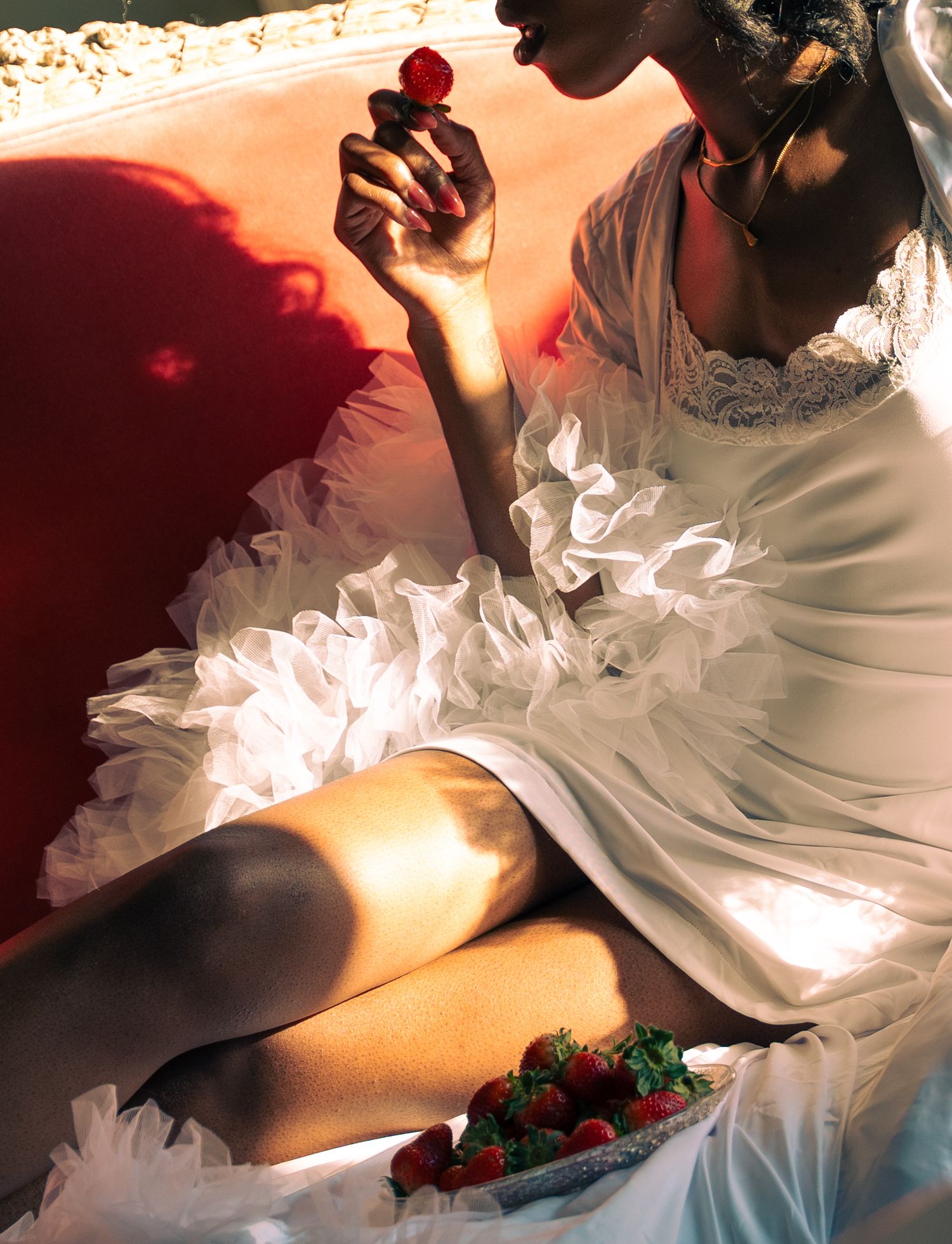 A Black bride in a white ruffled gown holds a red rose while seated near strawberries in soft natural light.
