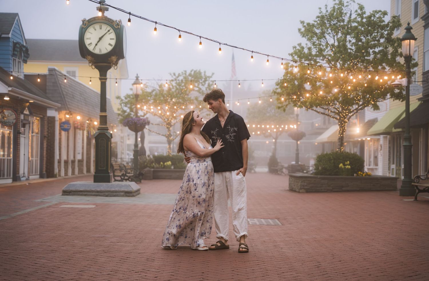 Romantic couple kissing near vintage clock under string lights in foggy town square.